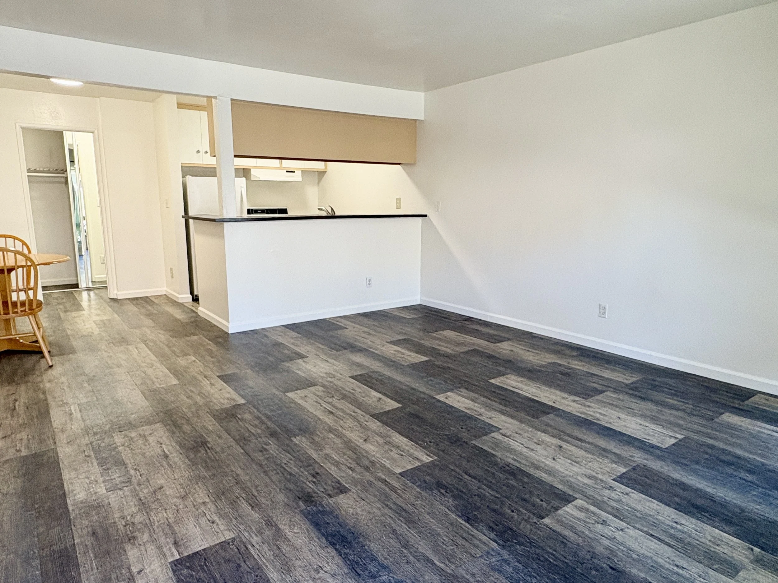 Empty living room with white walls, dark wood flooring, kitchen area with cabinets, and a dining table with chairs.