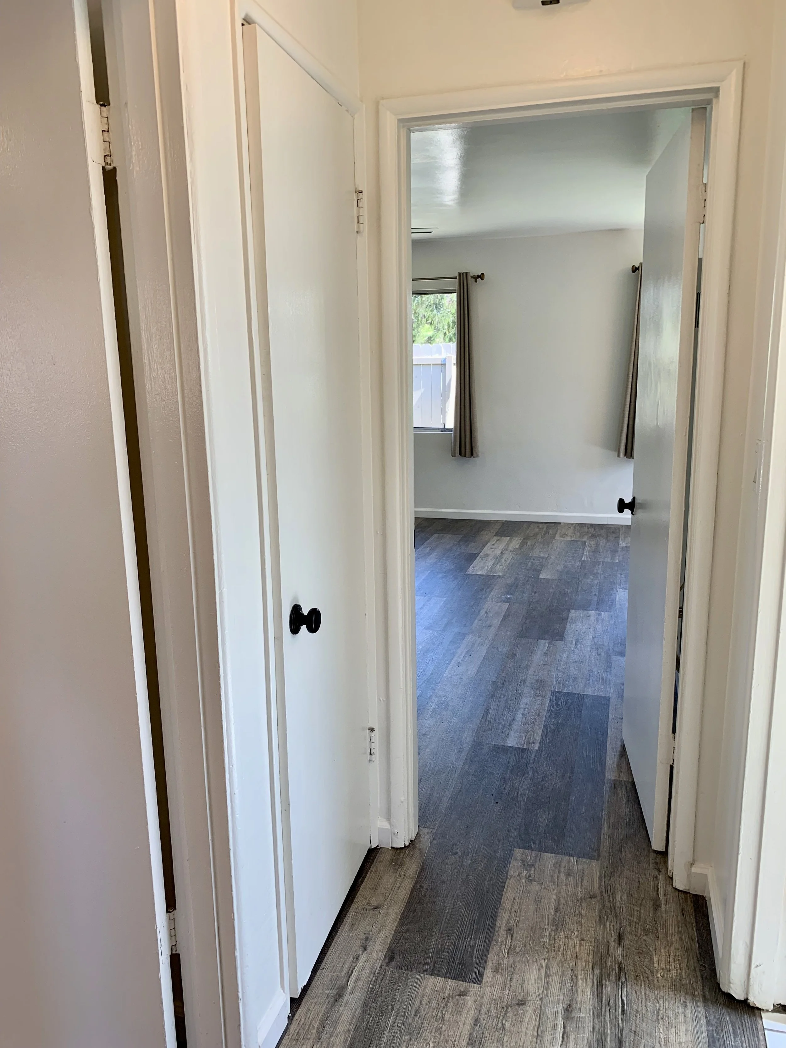 A hallway view with a partially open door leading to a room with a window and curtains, and a wooden floor.
