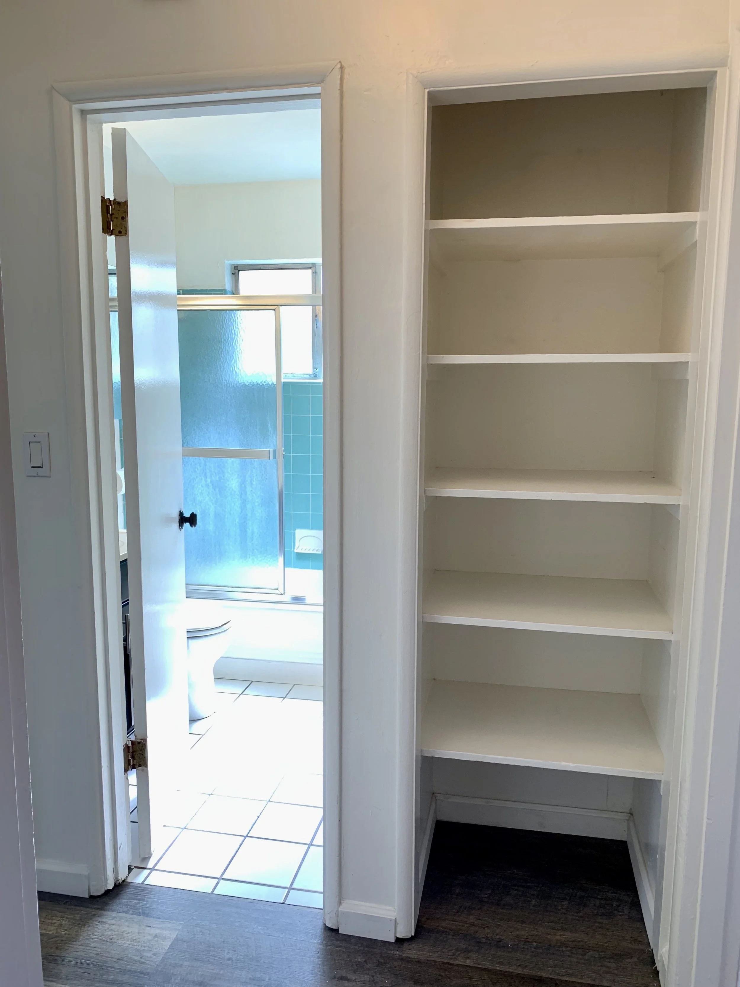 View of a bathroom looking through an open doorway with a small closet on the right and white-tiled floor in the bathroom, featuring a green tiled shower area with glass doors and a small window.