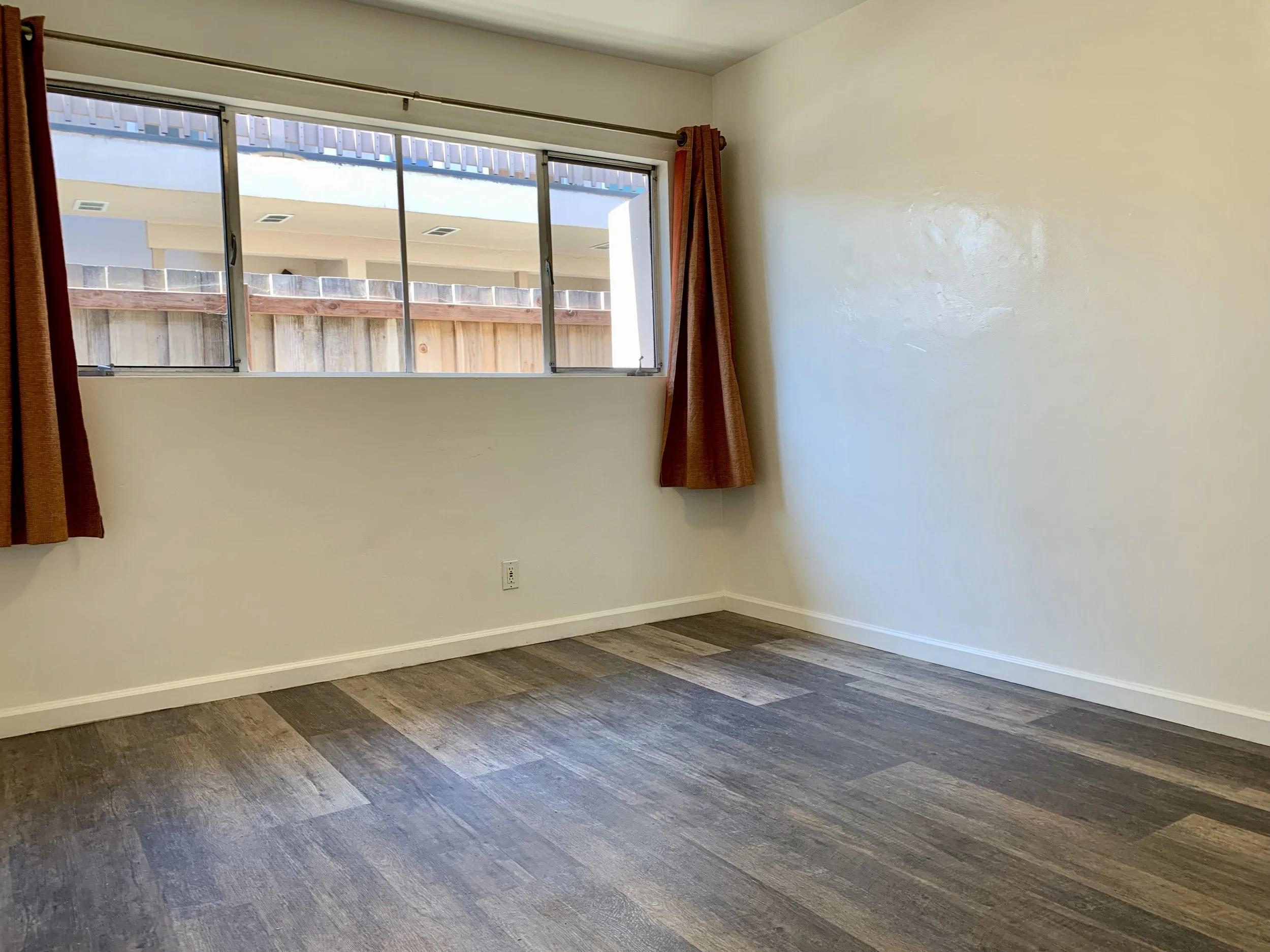 Empty room with hardwood floors, off-white walls, a window with curtains, and a view of a neighboring building and wooden fence outside.