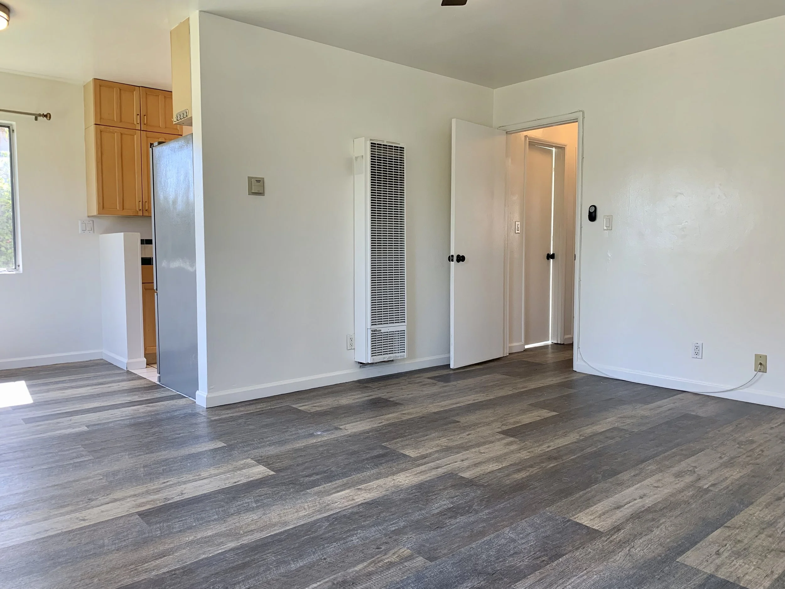 Empty room with white walls, gray wood flooring, a white wall heater, and an open white door leading to another room