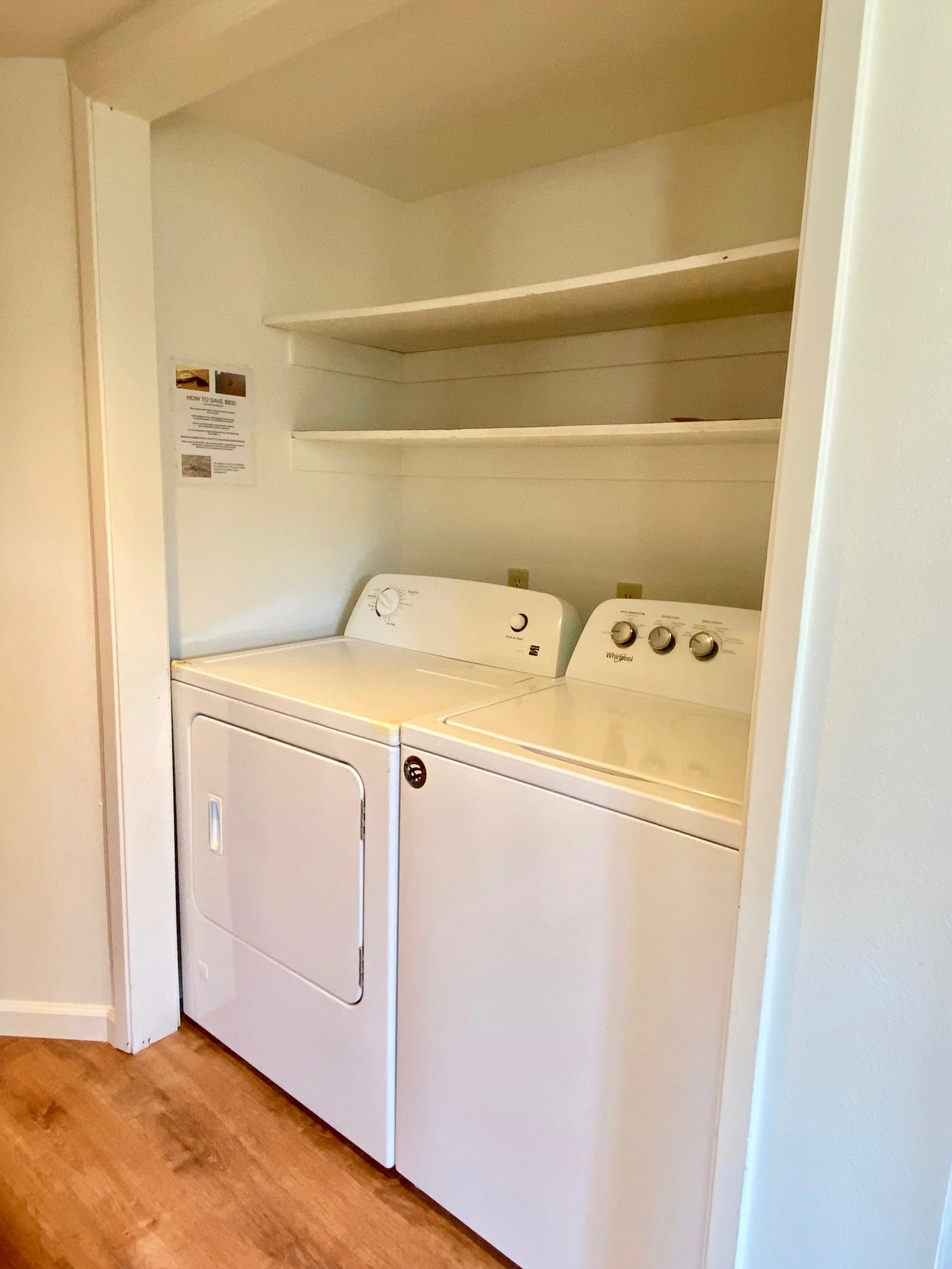 A laundry area with a white washing machine and dryer side by side beneath two empty white shelves, against a white wall with wood flooring.