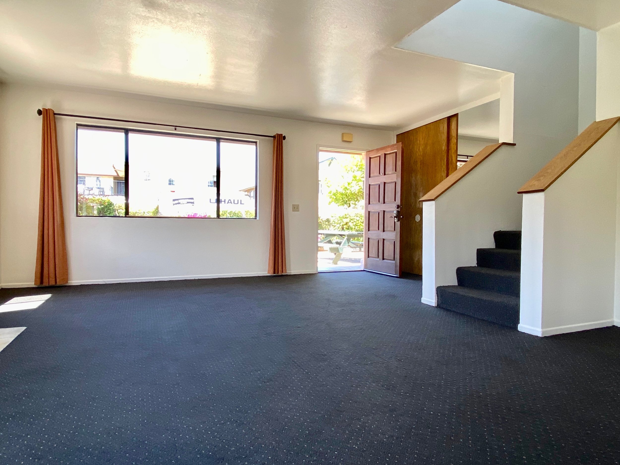 Living room with large window, open front door, staircase, dark carpet, sunlight coming through the window, and white walls.