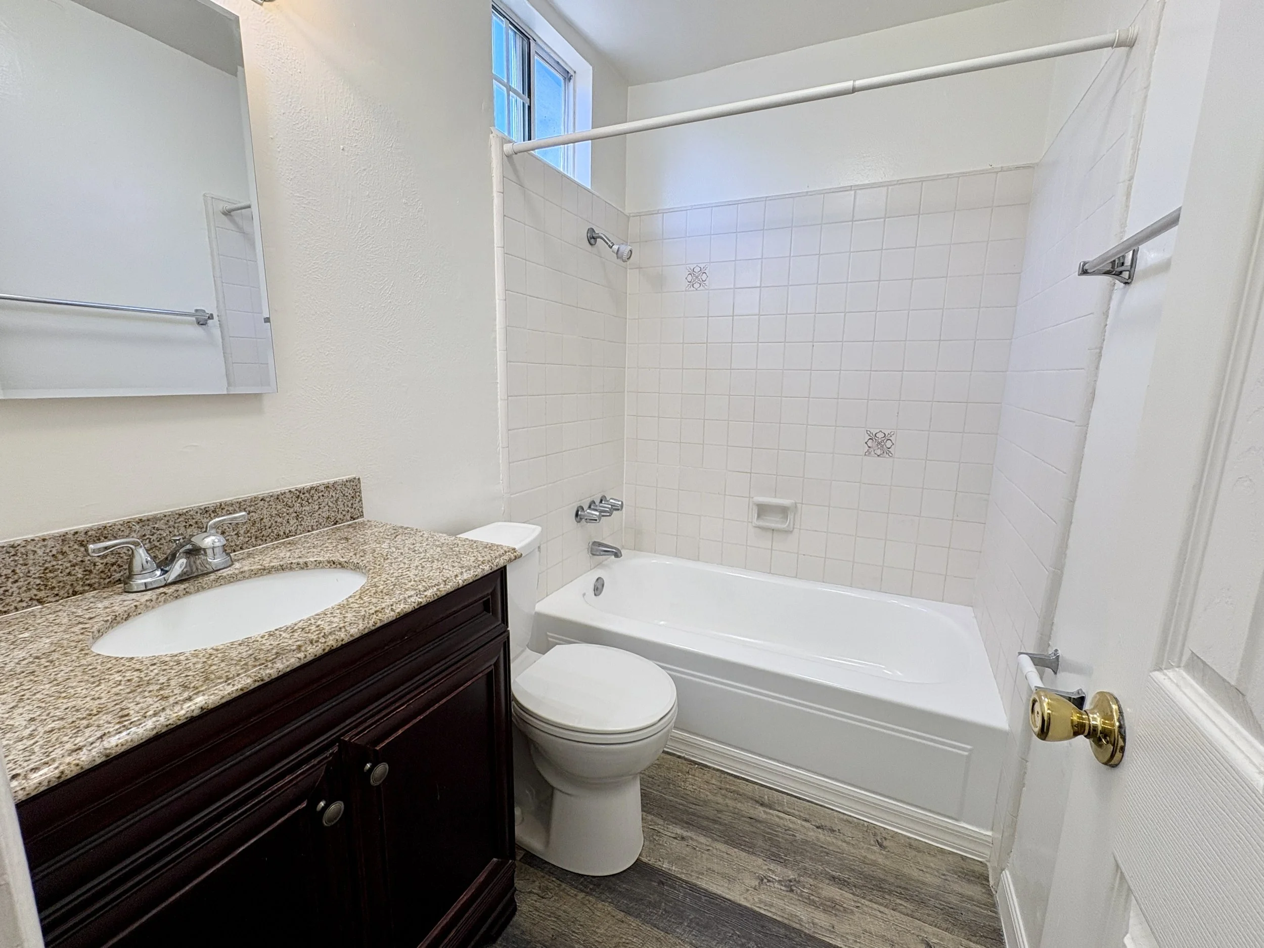 A bathroom with a granite countertop, a mirror, a toilet, a bathtub with a shower, a window, a towel bar, and a shower curtain rod.