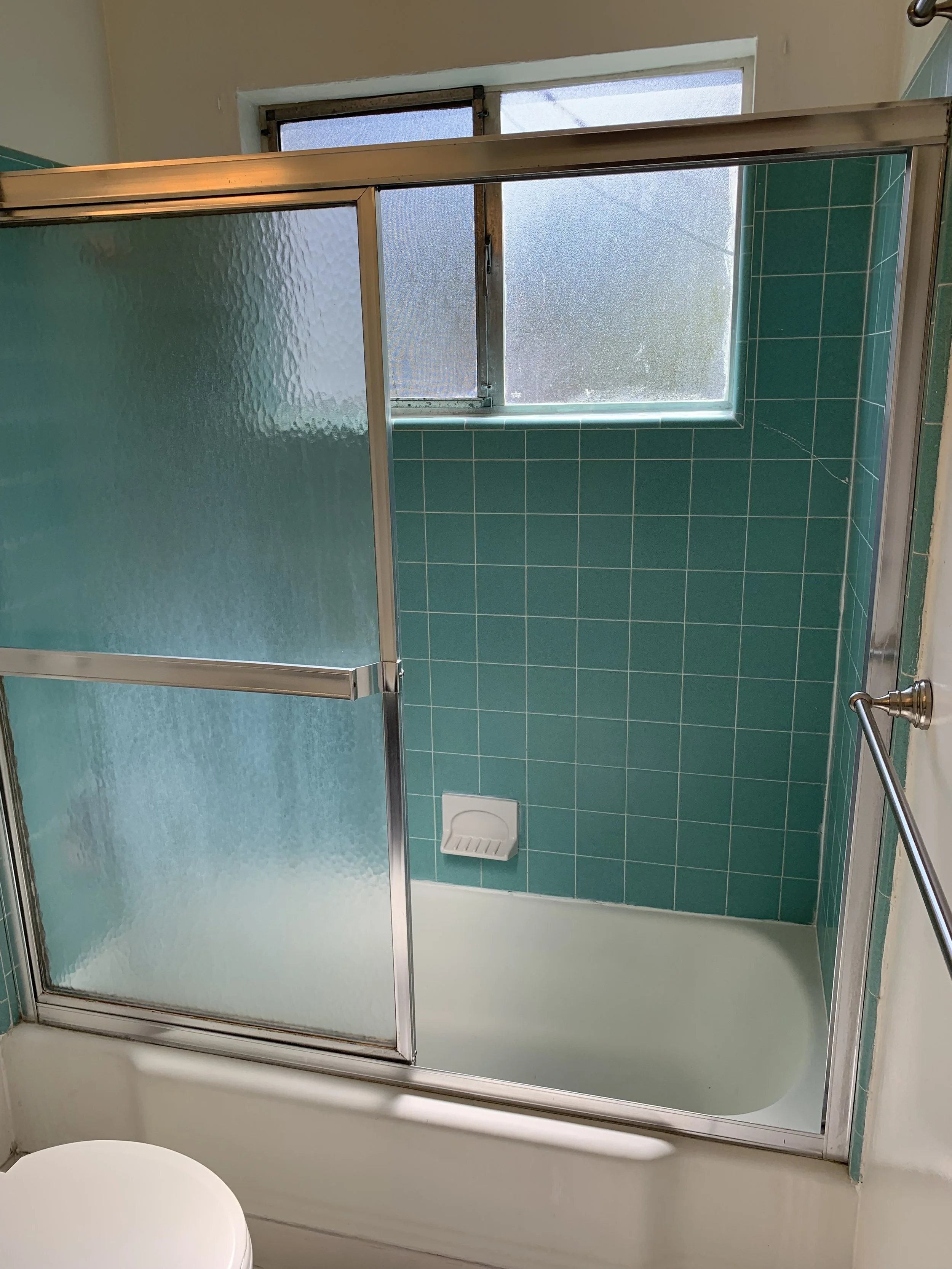 A bathroom with a shower stall that has green tiles, a frosted glass sliding door, a small soap dish, a frosted window, and a towel bar.