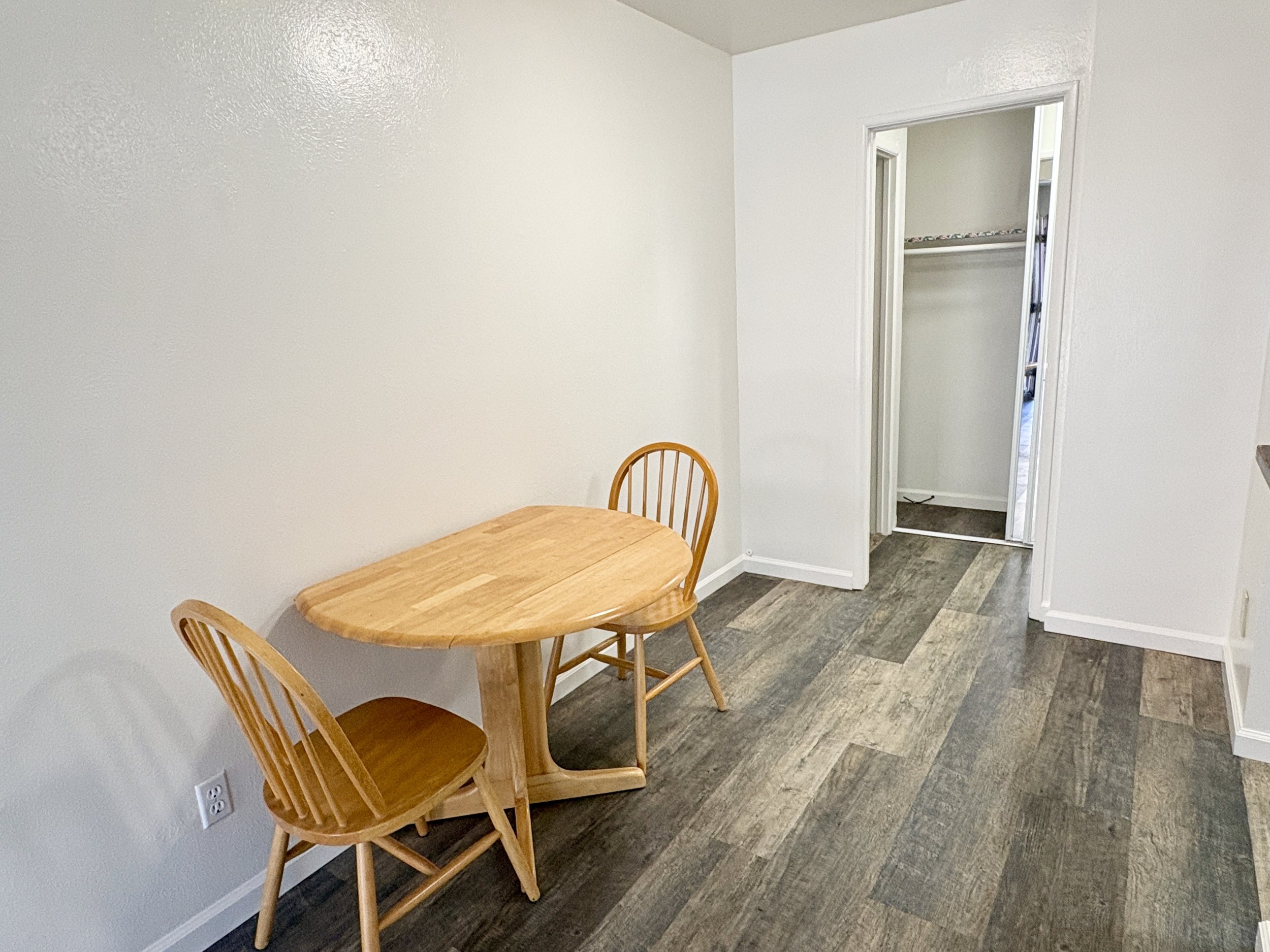 A corner of a room with a small wooden dining table and two matching chairs. An open closet door reveals empty shelves inside. The flooring is vinyl with a wood pattern, and the walls are painted white.