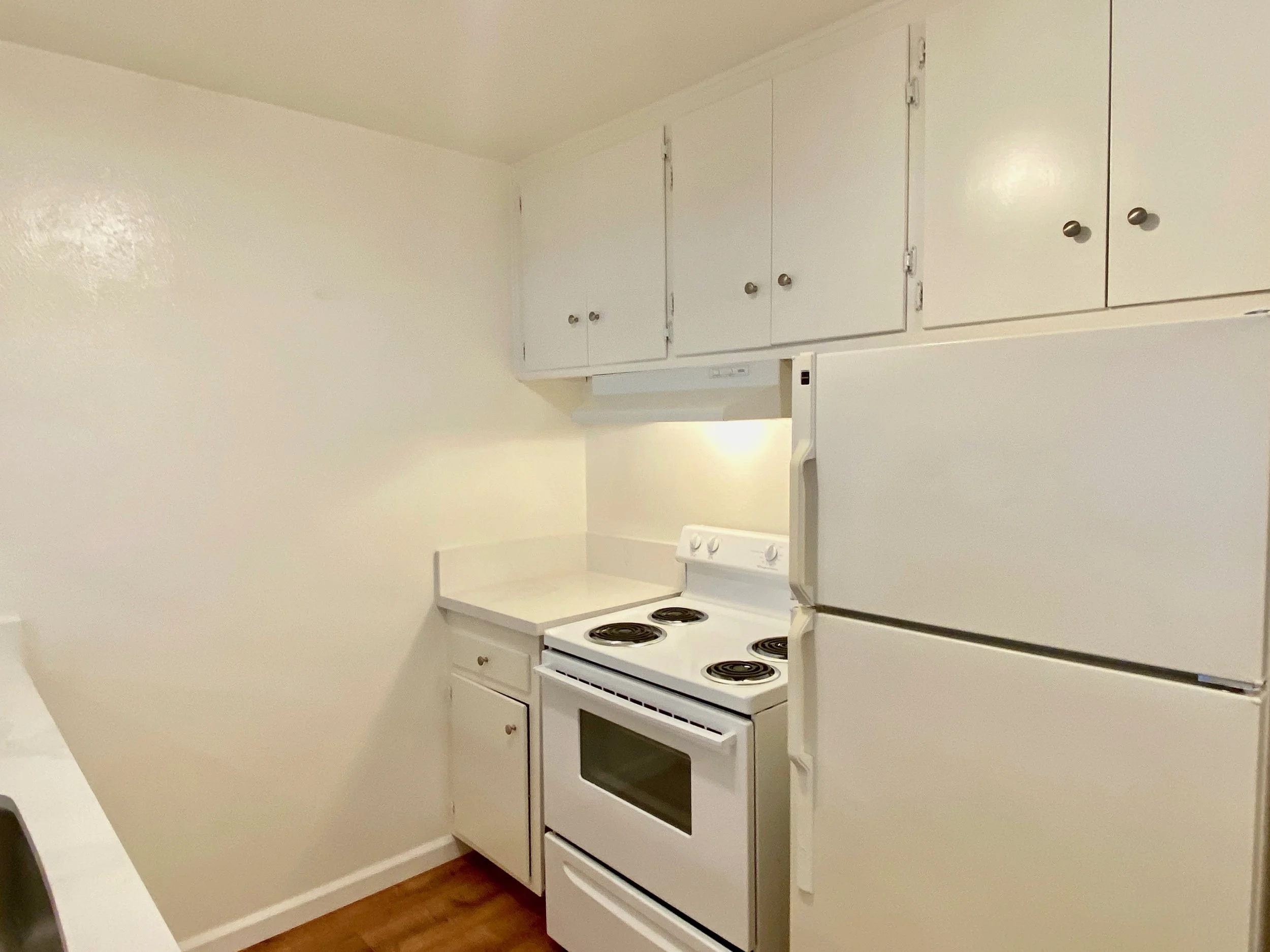 A small kitchen with white cabinets, a white stove with four coil burners, and a small white countertop with a cabinet underneath. The walls are painted white and the floor appears to be wood or wood-like material.