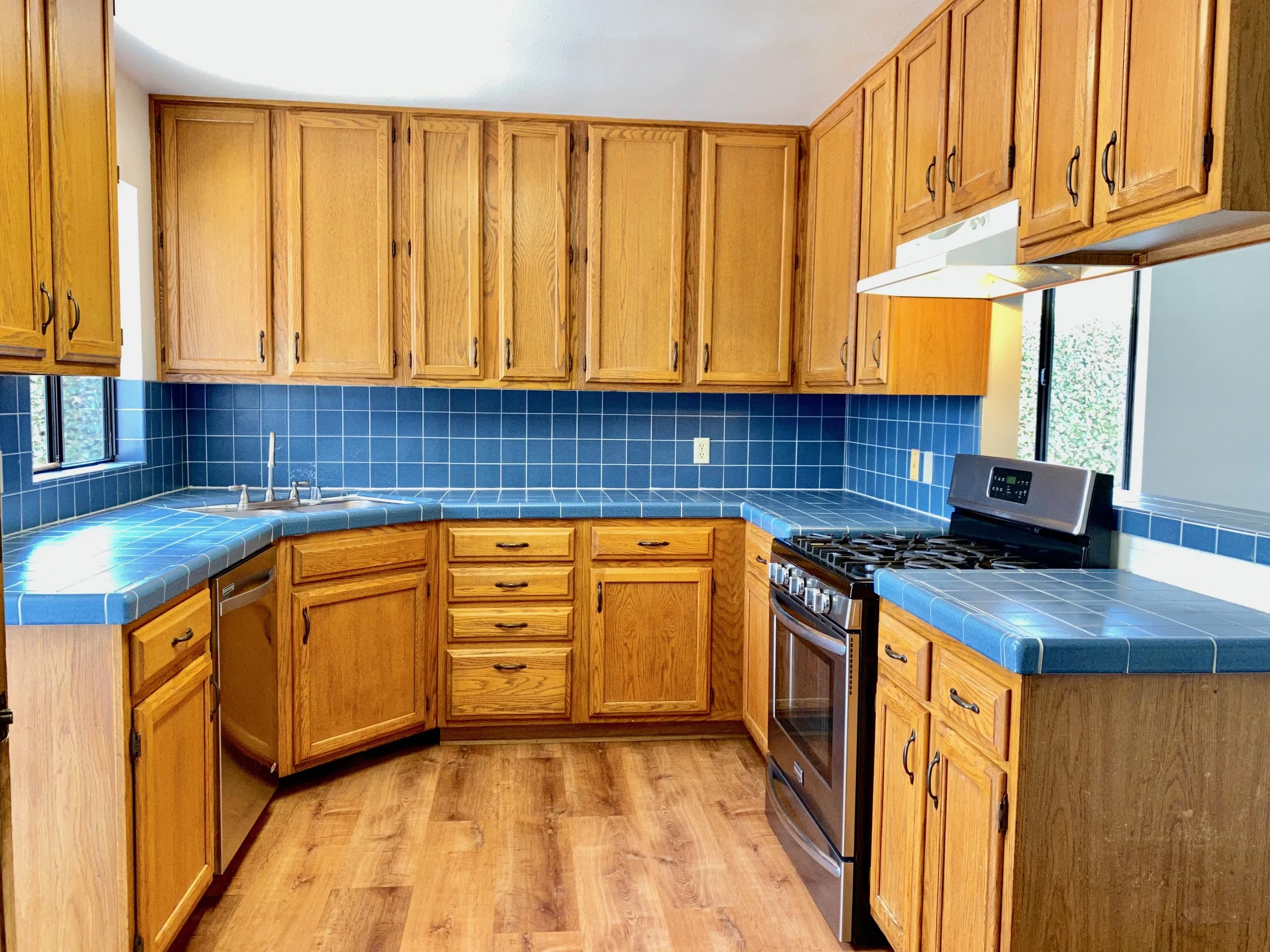 Kitchen with wooden cabinets, blue tiled countertops, a stainless steel stove, and a window.