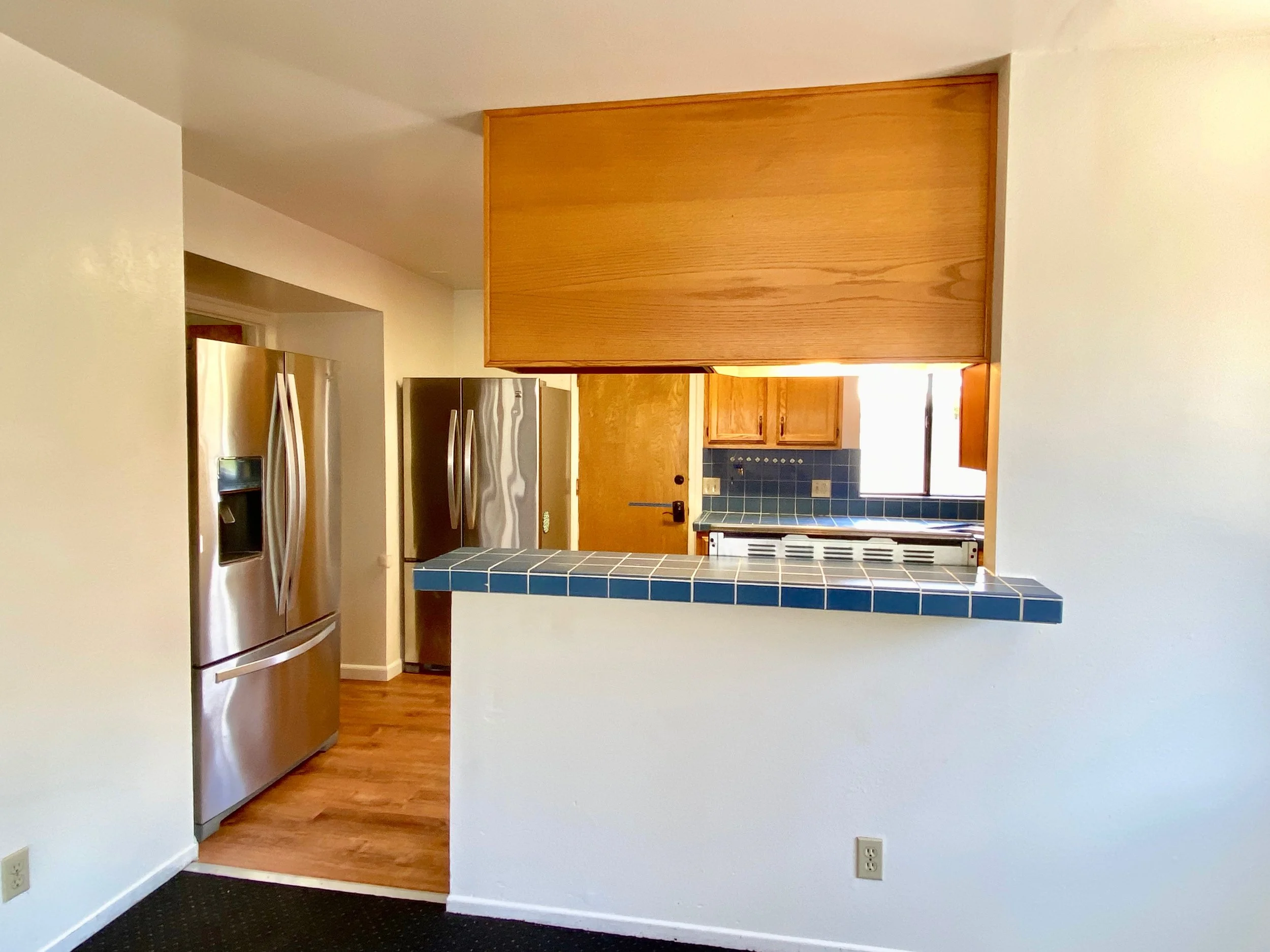 Interior view of a kitchen with a stainless steel refrigerator, wooden cabinets, and a blue tiled backsplash. Part of a hallway and a door are visible.