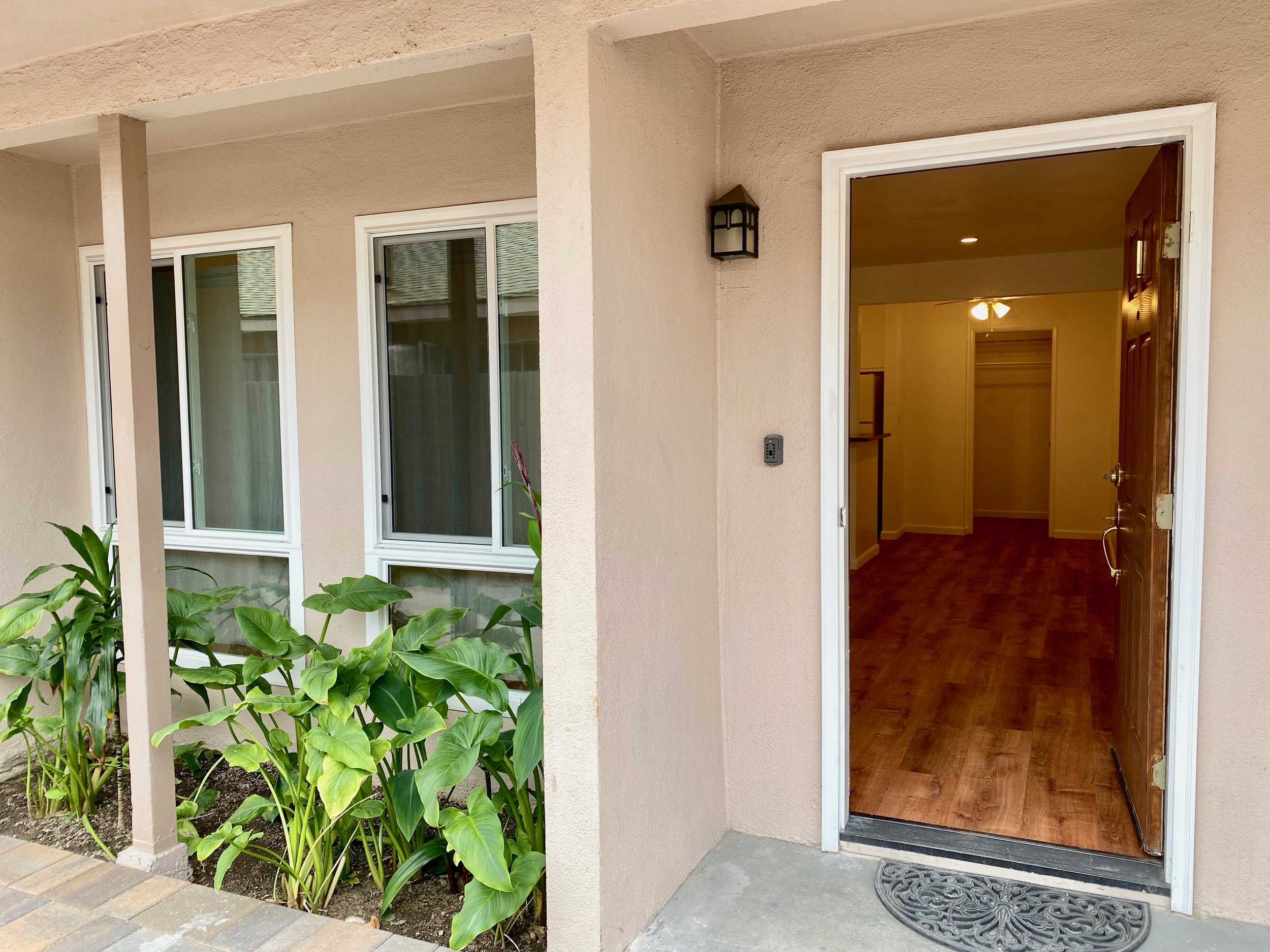 Close-up view of a house entrance with an open door leading to a room with wooden floors and ceiling fan, adjacent to a window with white frames and greenery outside.