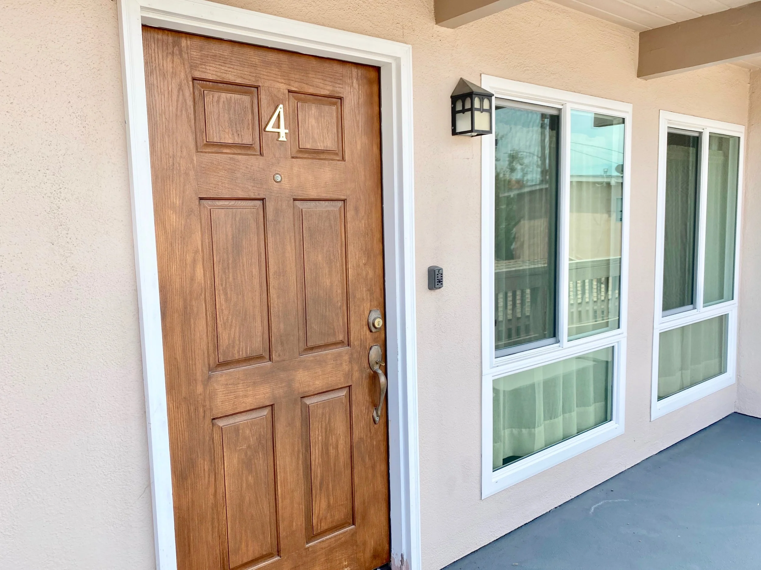 Front door of a house with a wooden door, a house number 4, and a black outdoor lantern. Next to the door are two windows with white frames and light-colored curtains.