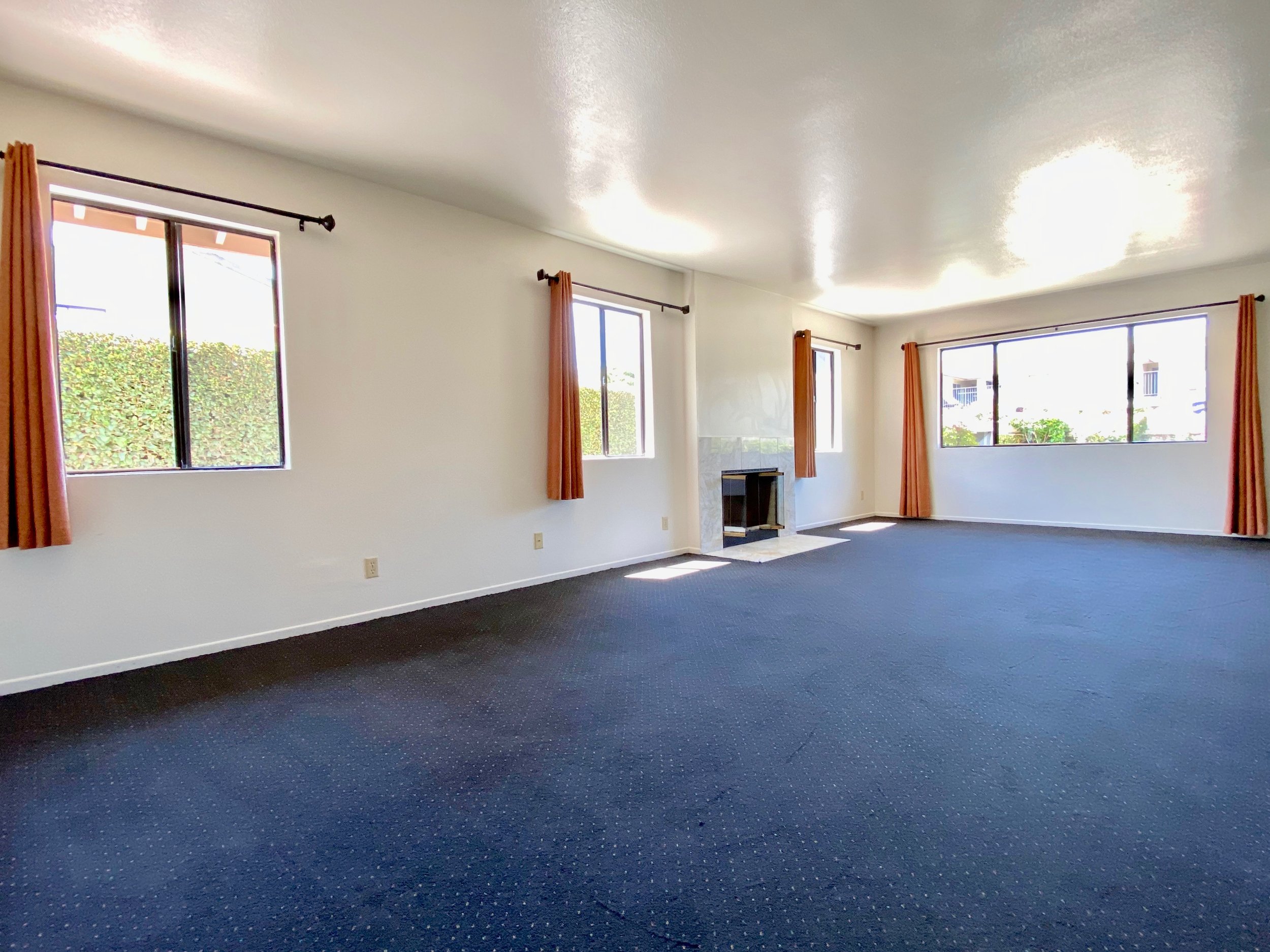 Empty living room with large windows, orange curtains, white walls, a fireplace, and a dark carpeted floor.