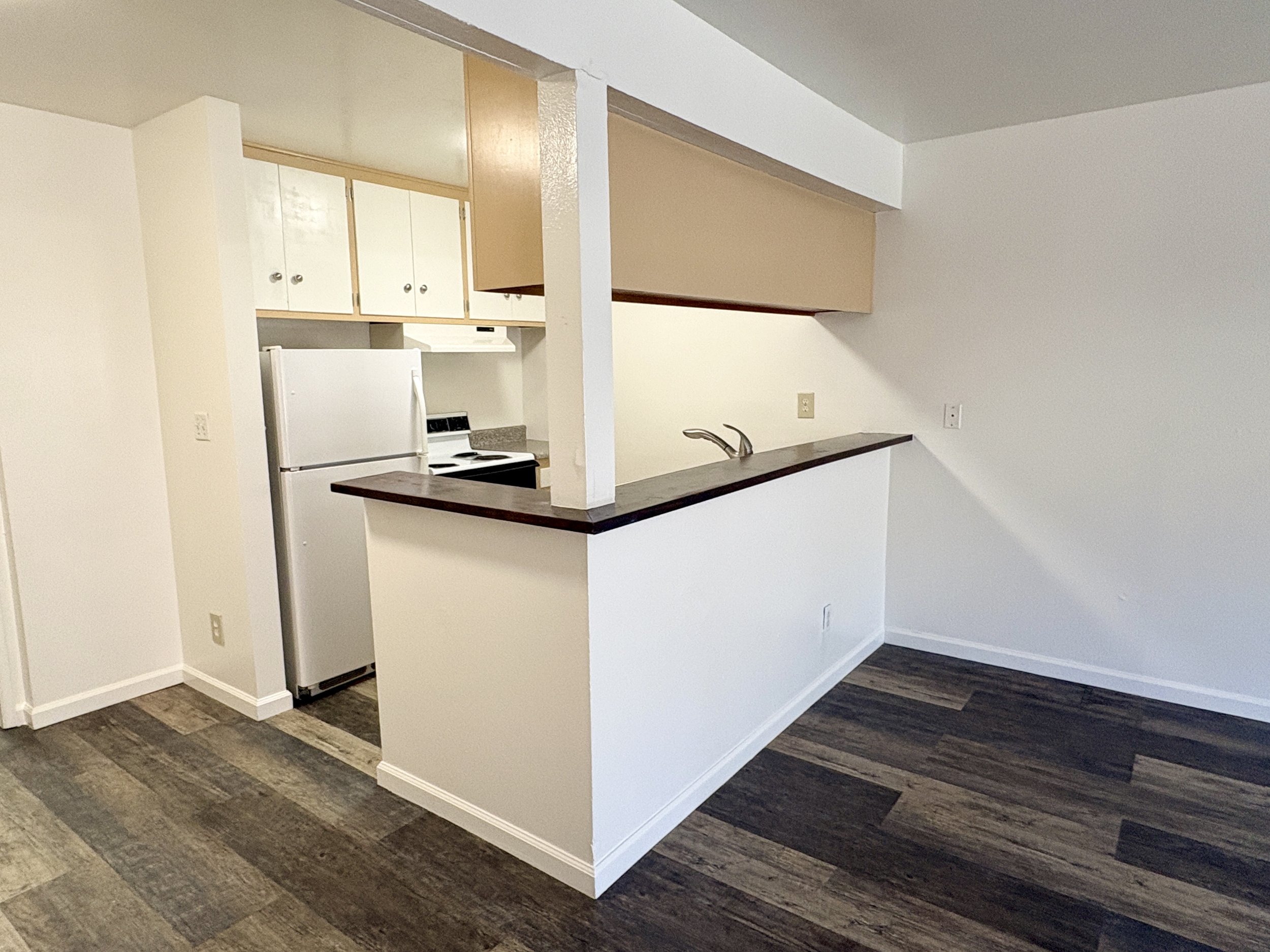 Kitchen with white refrigerator, beige upper cabinets, white wall, dark wood flooring, and a breakfast bar with a dark wood countertop.