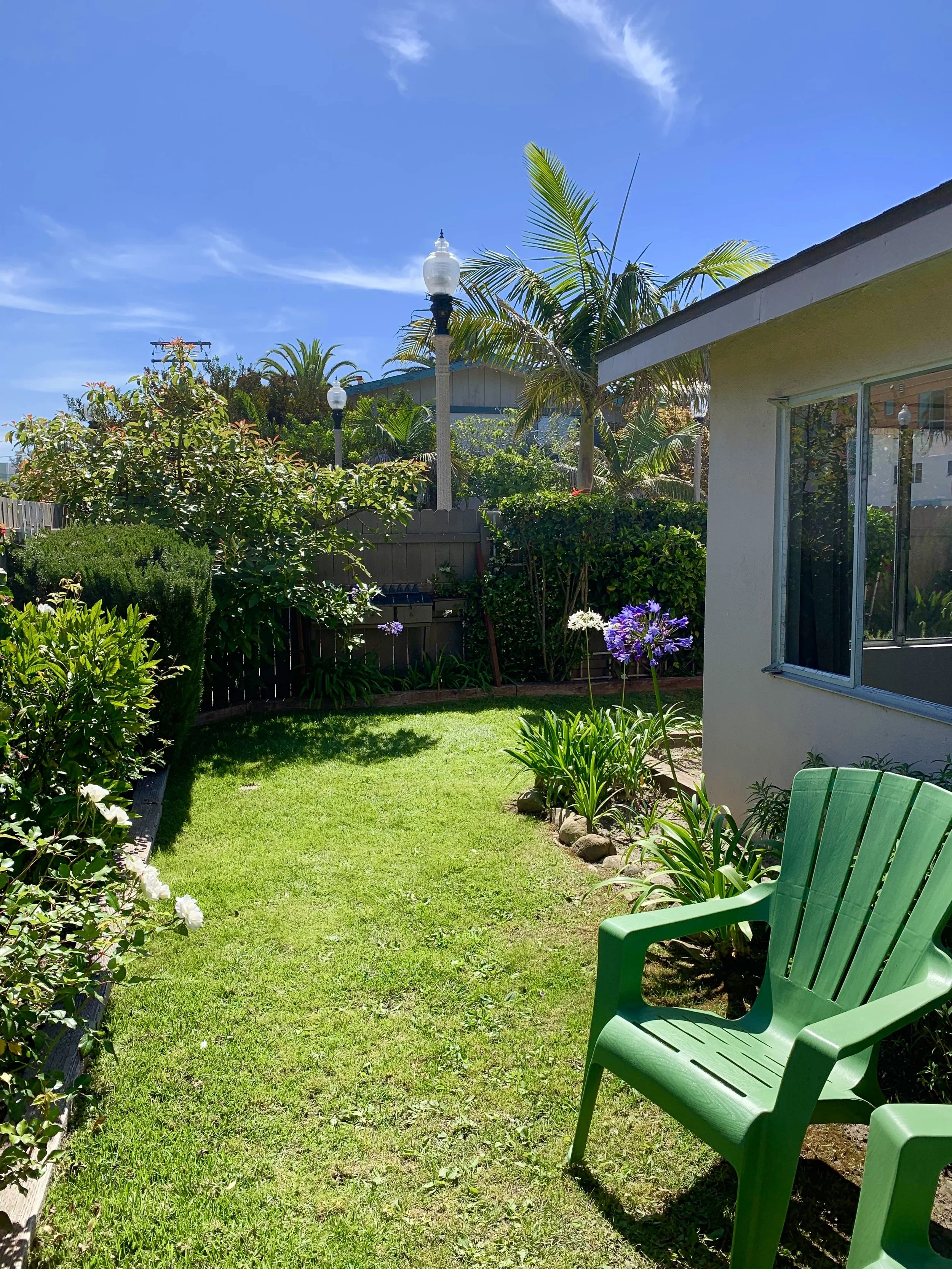 A backyard garden with a well-manicured lawn, surrounded by lush green plants and blooming flowers. There is a green Adirondack chair on the right side near a house with a  exterior and a large window. Tall palm trees, lamp posts, and a wooden fence 