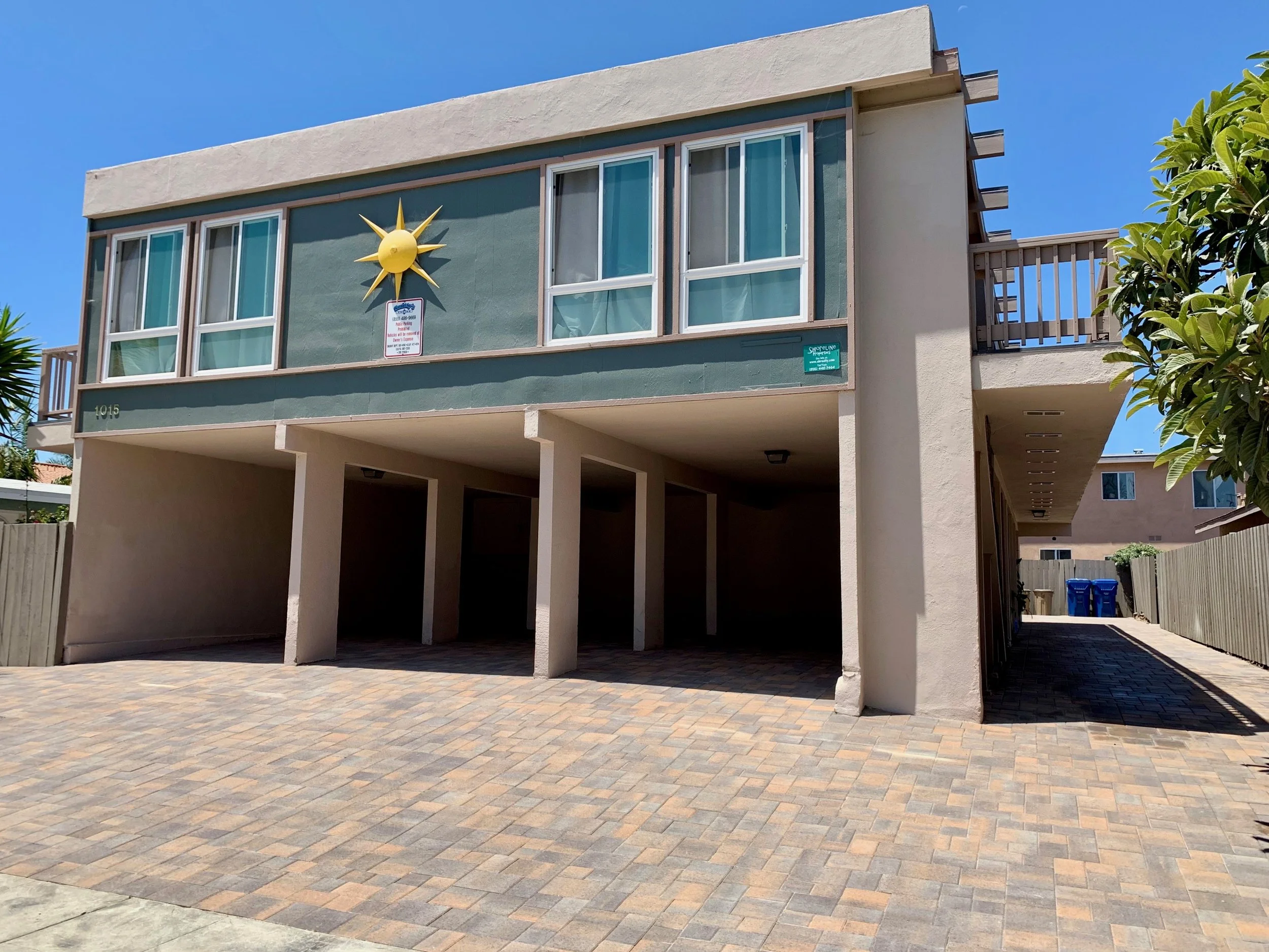 A multi-story residential building with a green exterior and white trim, featuring large windows, a small balcony, and a decorative sun sculpture on the front. The building is supported by columns and has a brick-paved area underneath, with blue recy