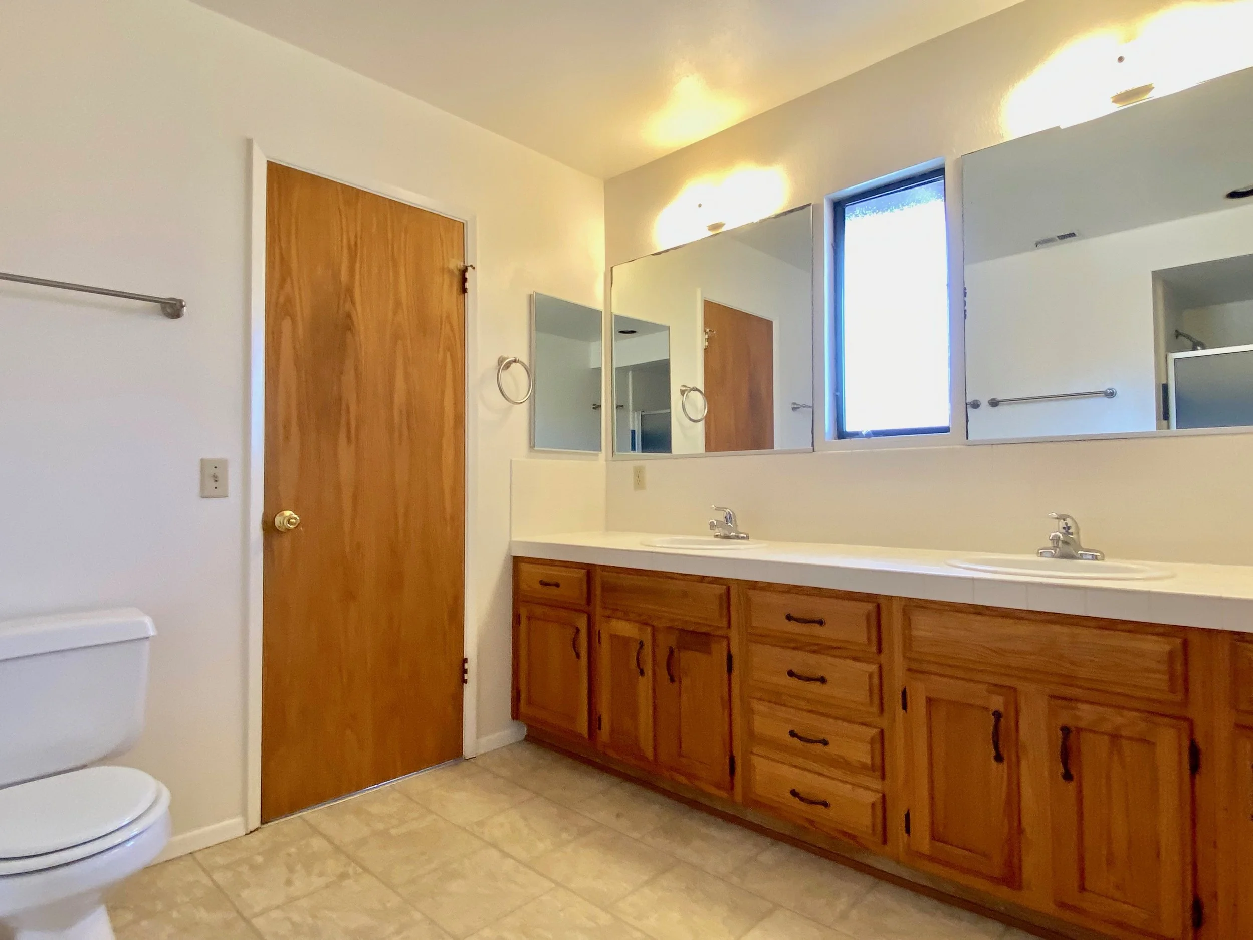 A bathroom with a double sink vanity with wooden cabinets, a large mirror, two towel rings, a window, a door, a toilet, and a shower in the background.
