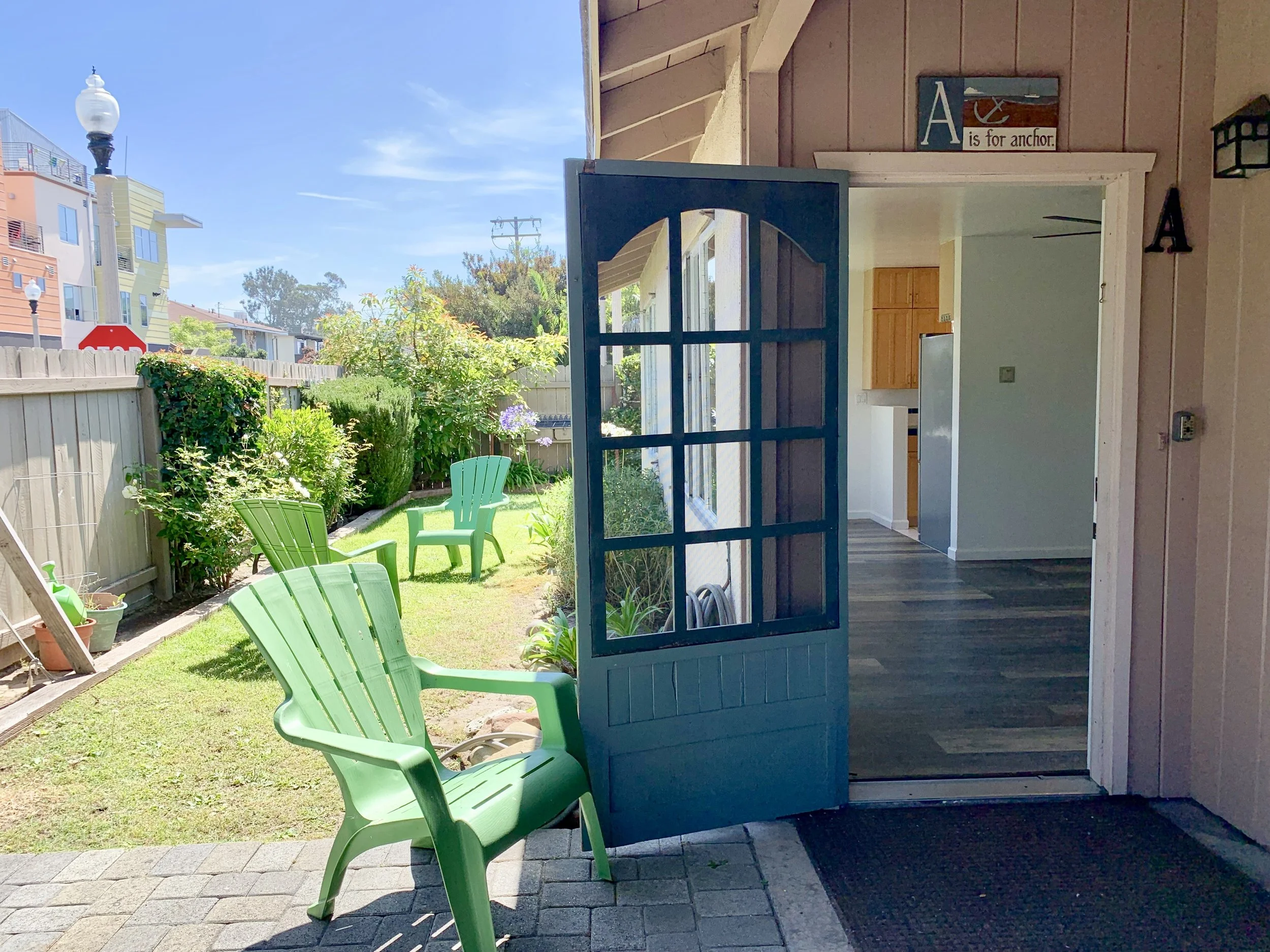 View of an apartment where the door is open. To the left of the door is a backyard with green chairs, plants, a wooden fence, and colorful modern apartment buildings in the background. The sky is clear and blue.
