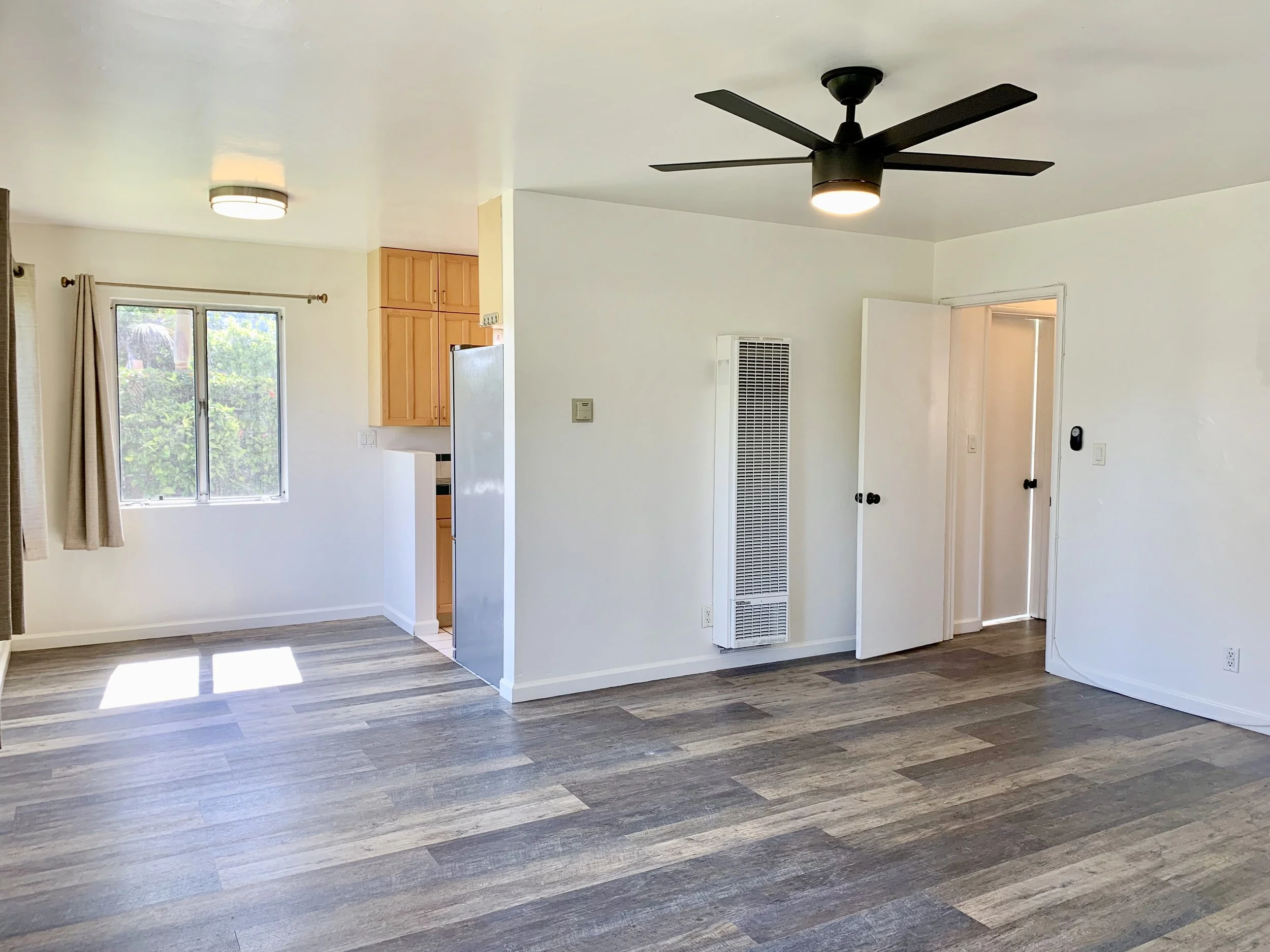 Living room with a window, beige curtains, a ceiling light, a modern black ceiling fan with light, wood flooring, white walls, and a doorway.