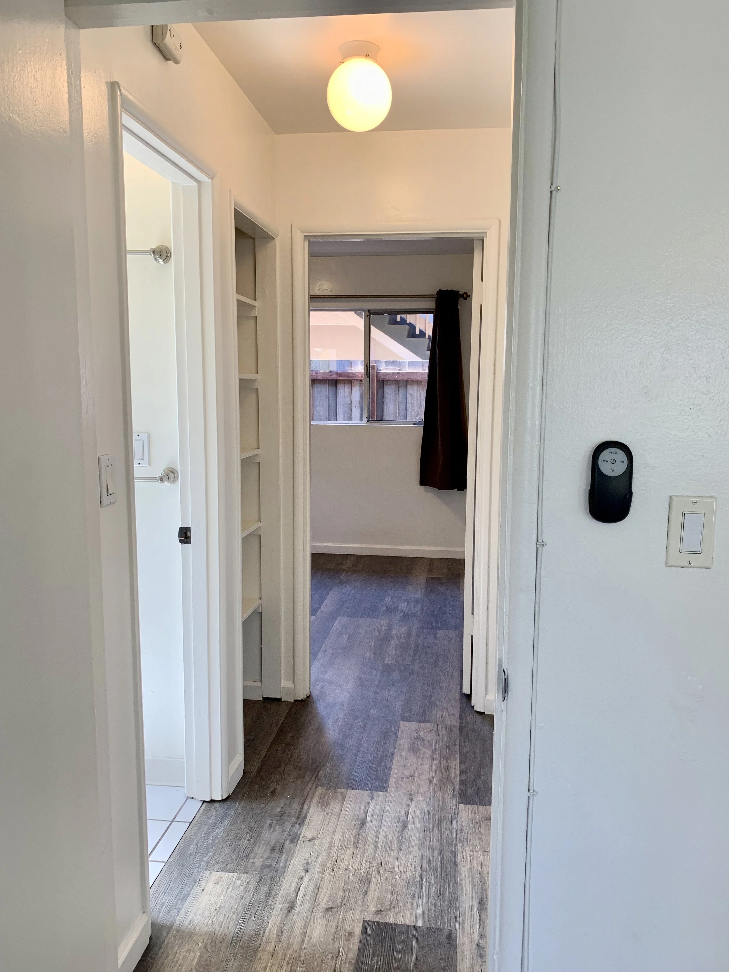 Hallway leading to a room with a window and dark curtains, hardwood flooring, and a ceiling light.