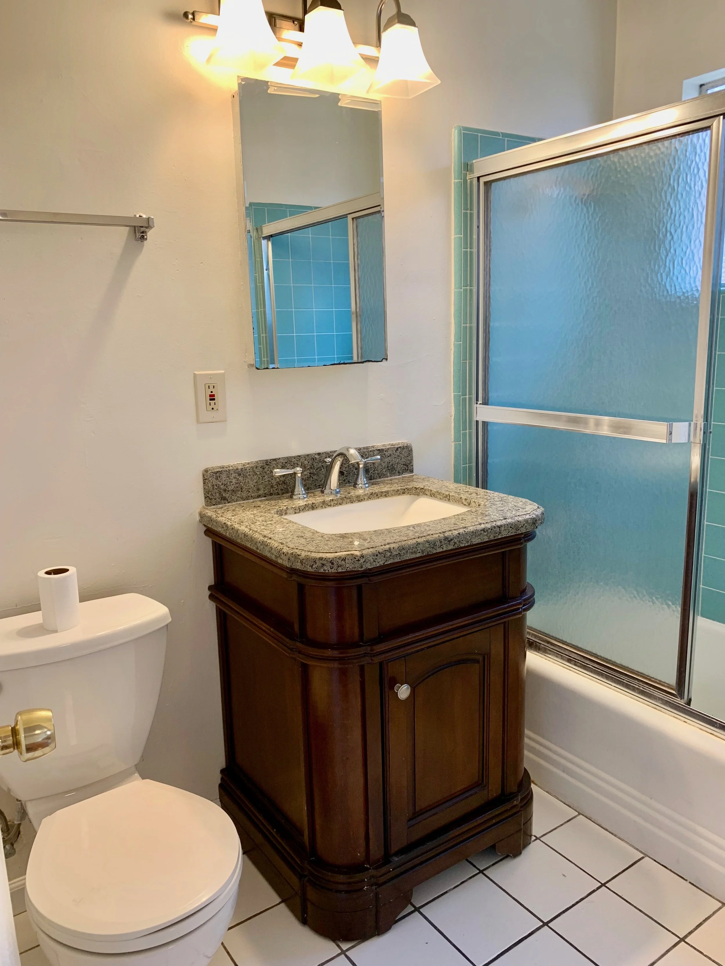 A bathroom with a white toilet, a granite countertop vanity with a dark wood base, a mirror, a light fixture with three lamps, a frosted glass shower door, green tiled wall inside the shower, and white floor tiles.
