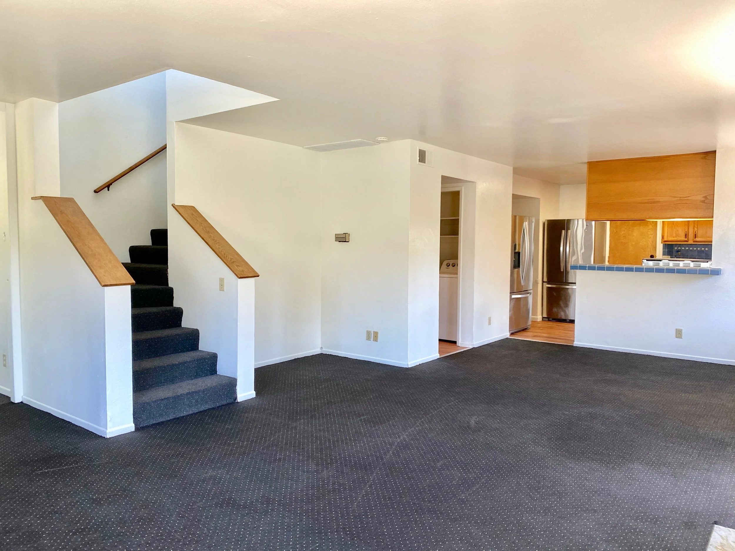 Interior view of a living space with black carpet, stairs with wooden handrails, and a doorway leading to a laundry room. In the background, part of a kitchen with stainless steel refrigerator and wooden cabinets is visible. The walls and ceiling are