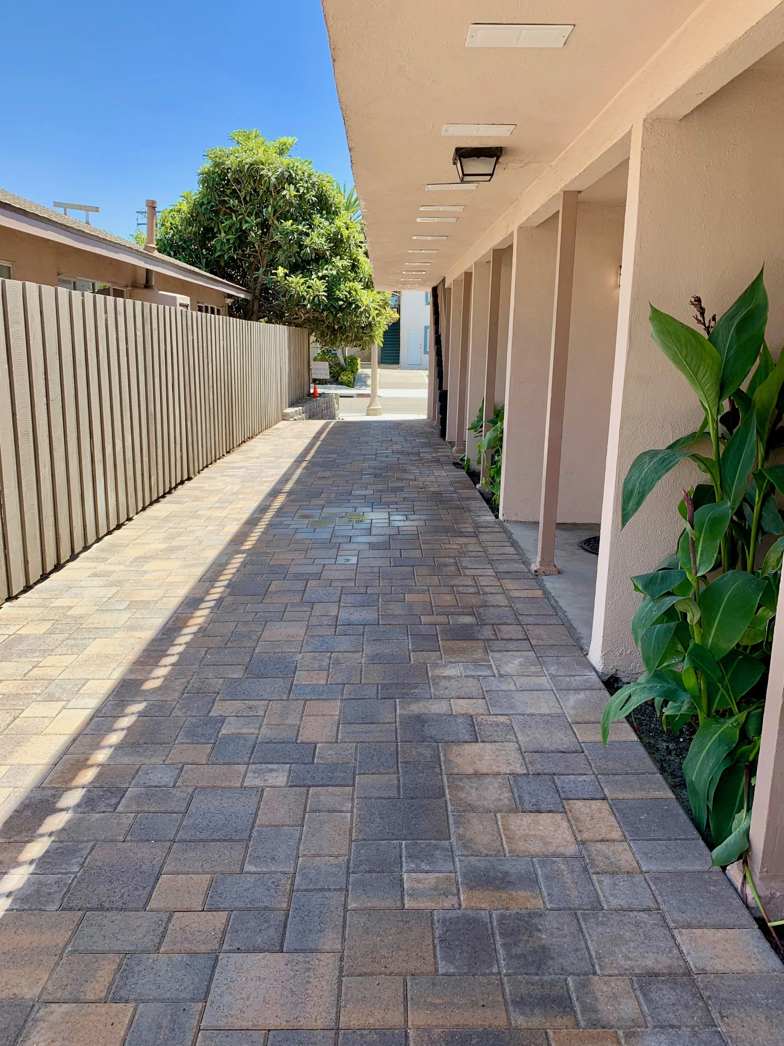 Exterior driveway with brick pavers, beige building to the right, wooden fence to the left, green trees, a lamppost at the end of the walkway, and a blue sky.
