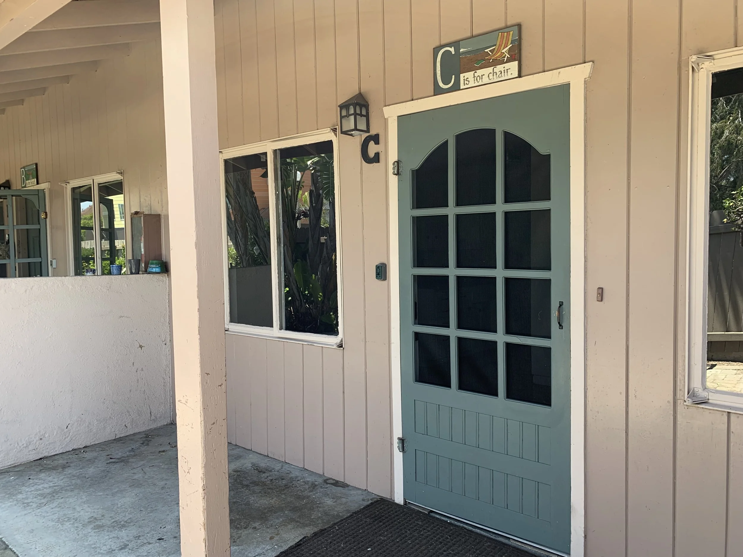 A front porch with a teal door, a small window with white trim, a sign that says 'C is for chair' with a beach chair illustration, a wall-mounted lantern light.