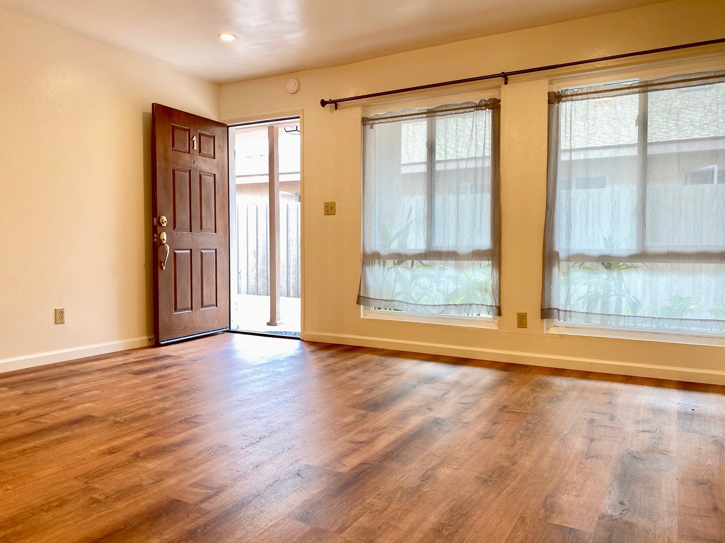 An empty room with wooden flooring, cream-colored walls, a door open to the outside, and large windows with sheer curtains.