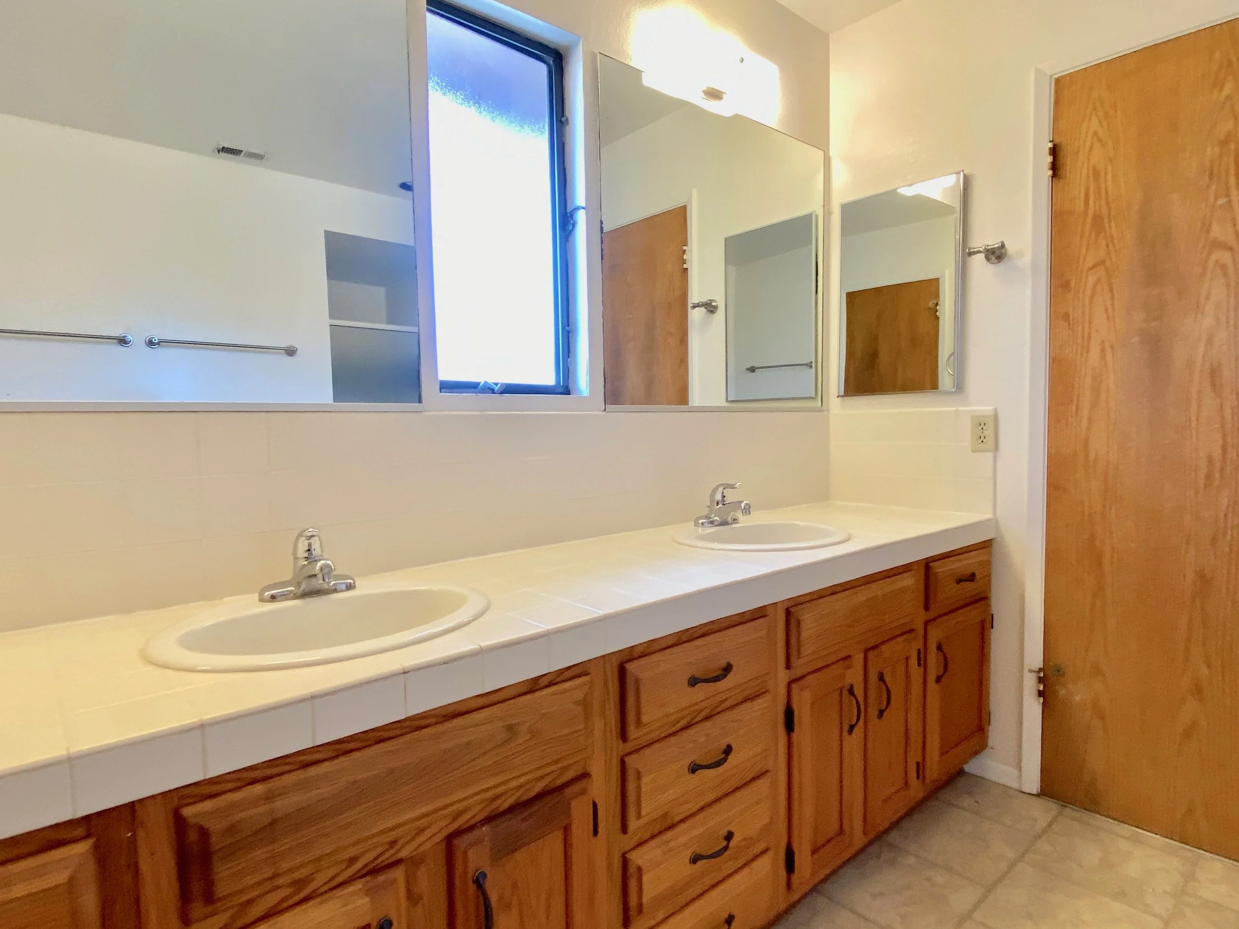 Bathroom vanity with dual sinks, wooden cabinets, large mirror, window, and wooden door.