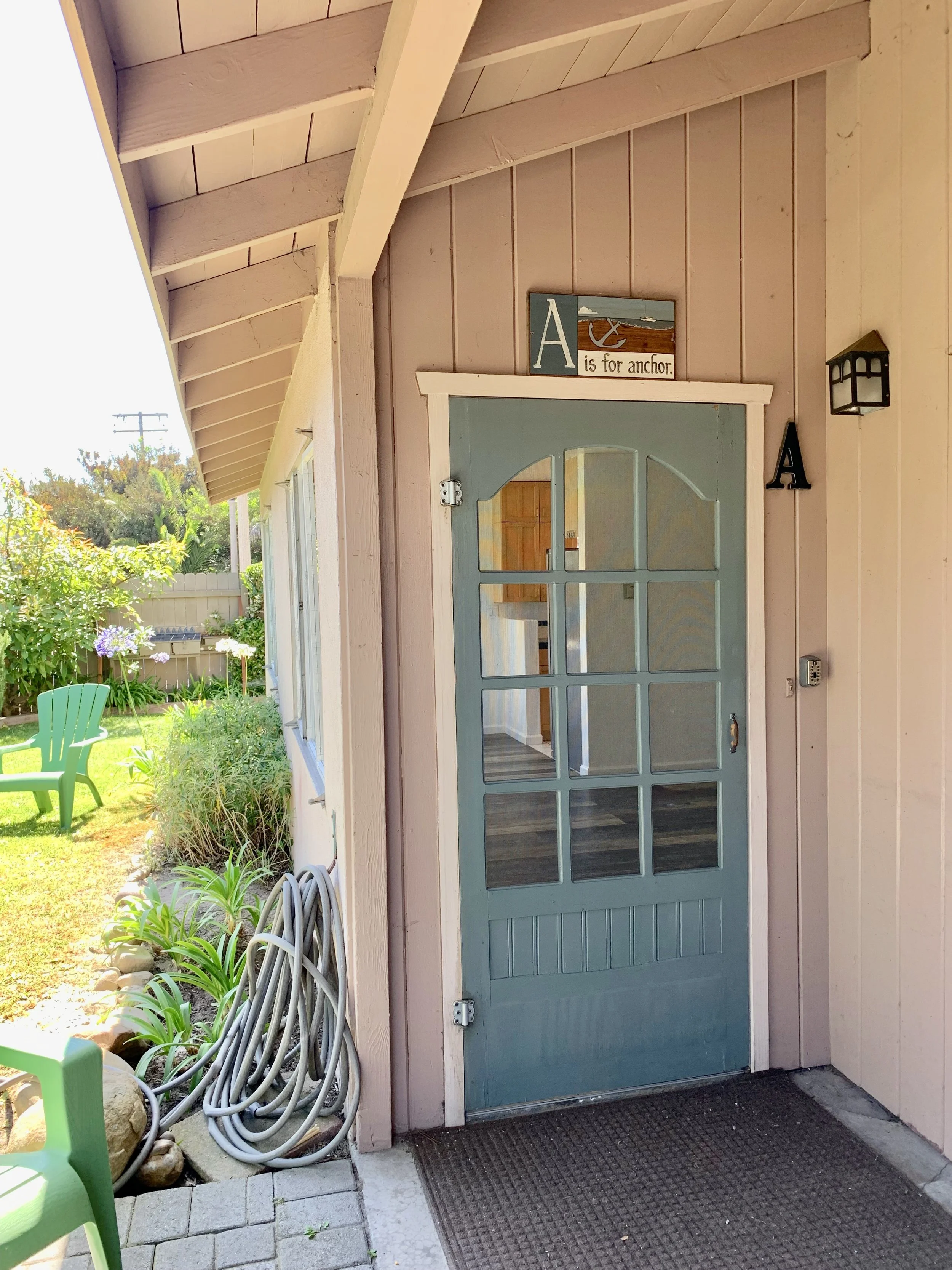 Exterior view of a house entrance with a light blue door, a sign above it that says "A is for anchor," a small porch, and garden with bushes and flowers. coiled hoses are on the side.