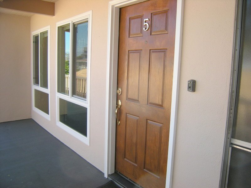 Close-up of a brown front door with the number 5, a door handle, and a lock, next to a small keypad, with windows to the left and a beige wall.