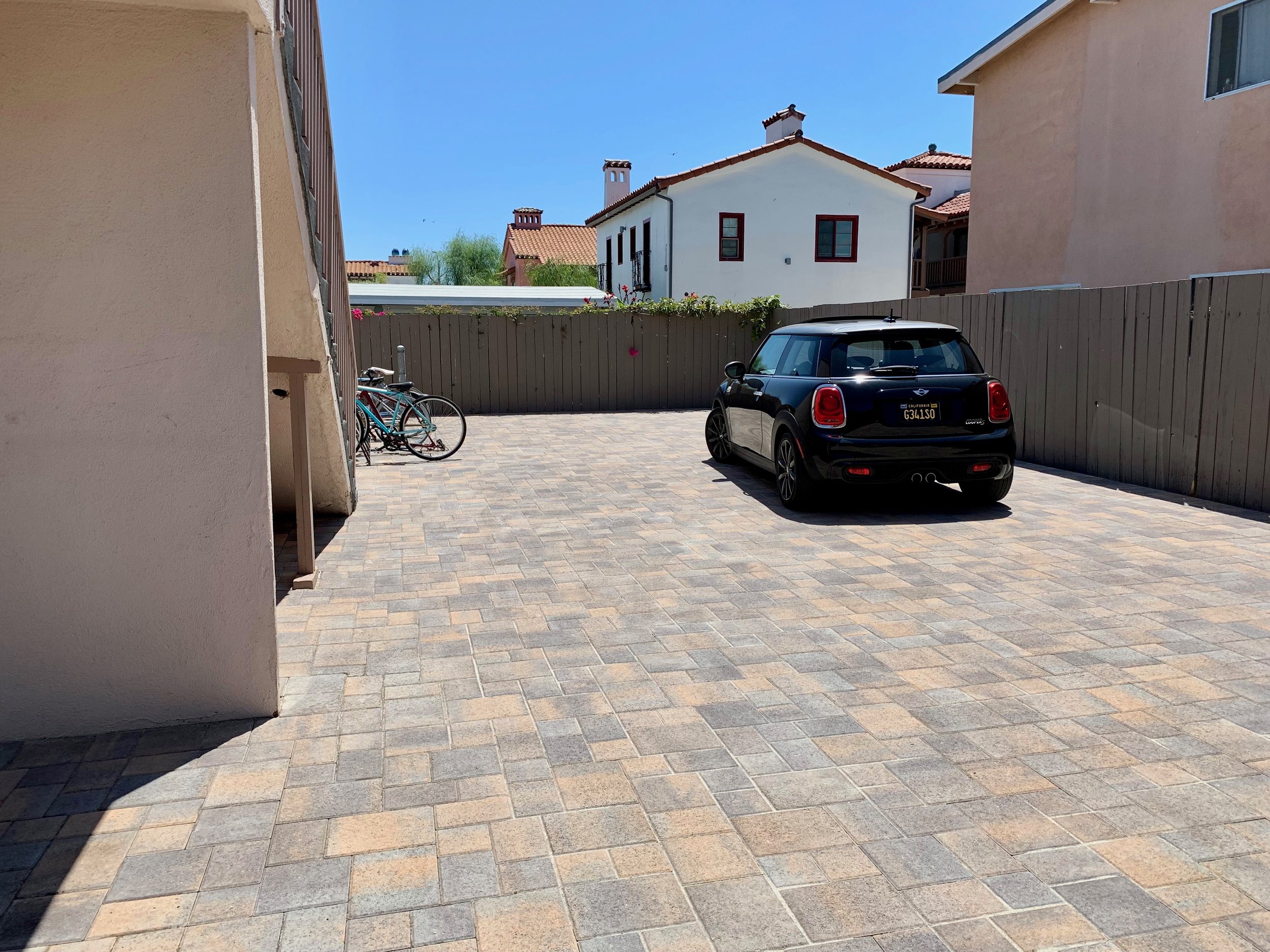 A sunny back driveway with brick paving, a black Mini Cooper car, two bicycles, a wooden fence with flowers, and houses with red-tiled roofs and chimneys in the background.