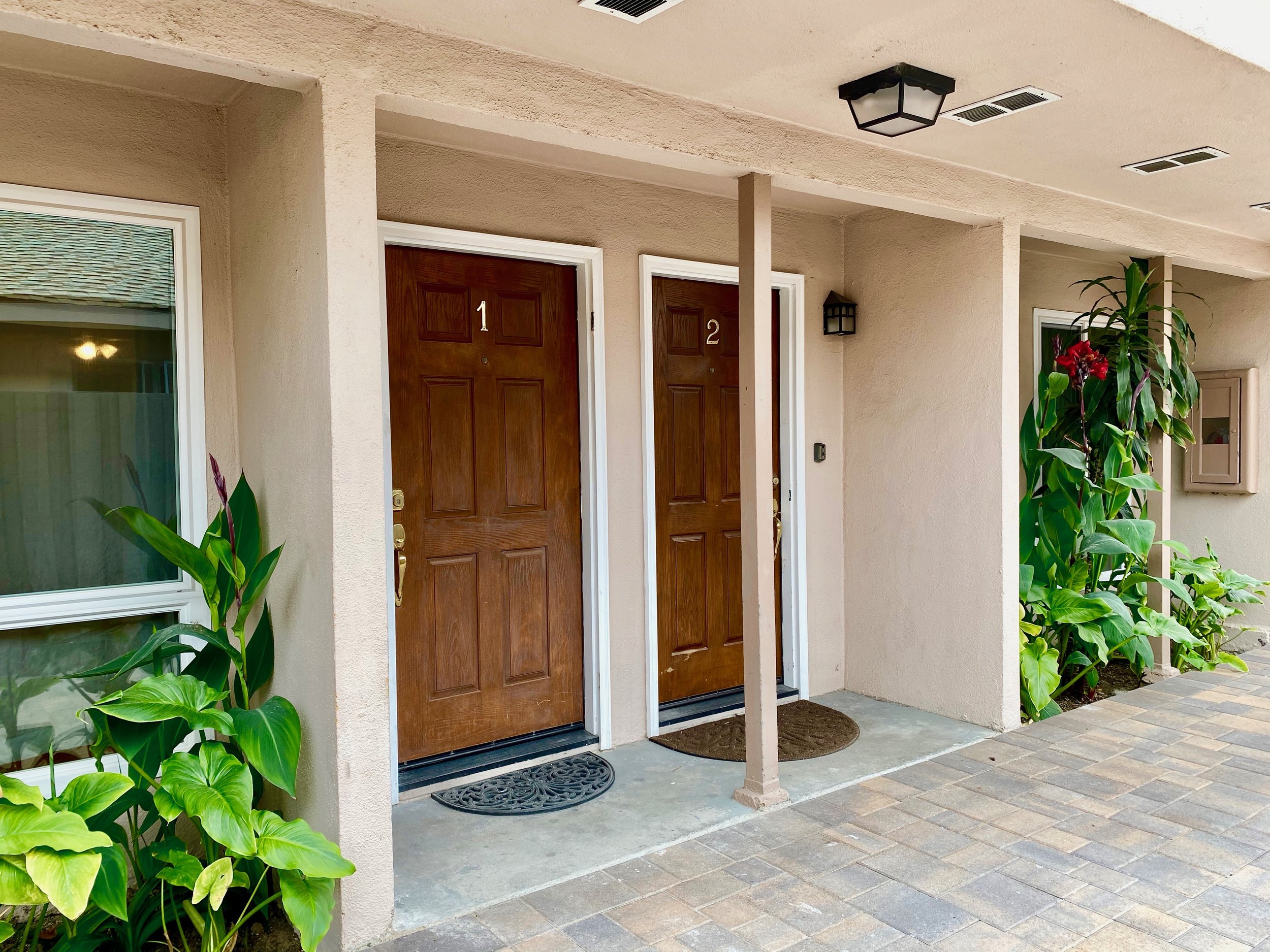 Front door of a residential building with two brown doors numbered 1 and 2, a planter area with green plants,  a ceiling light fixture, and a paved walkway.