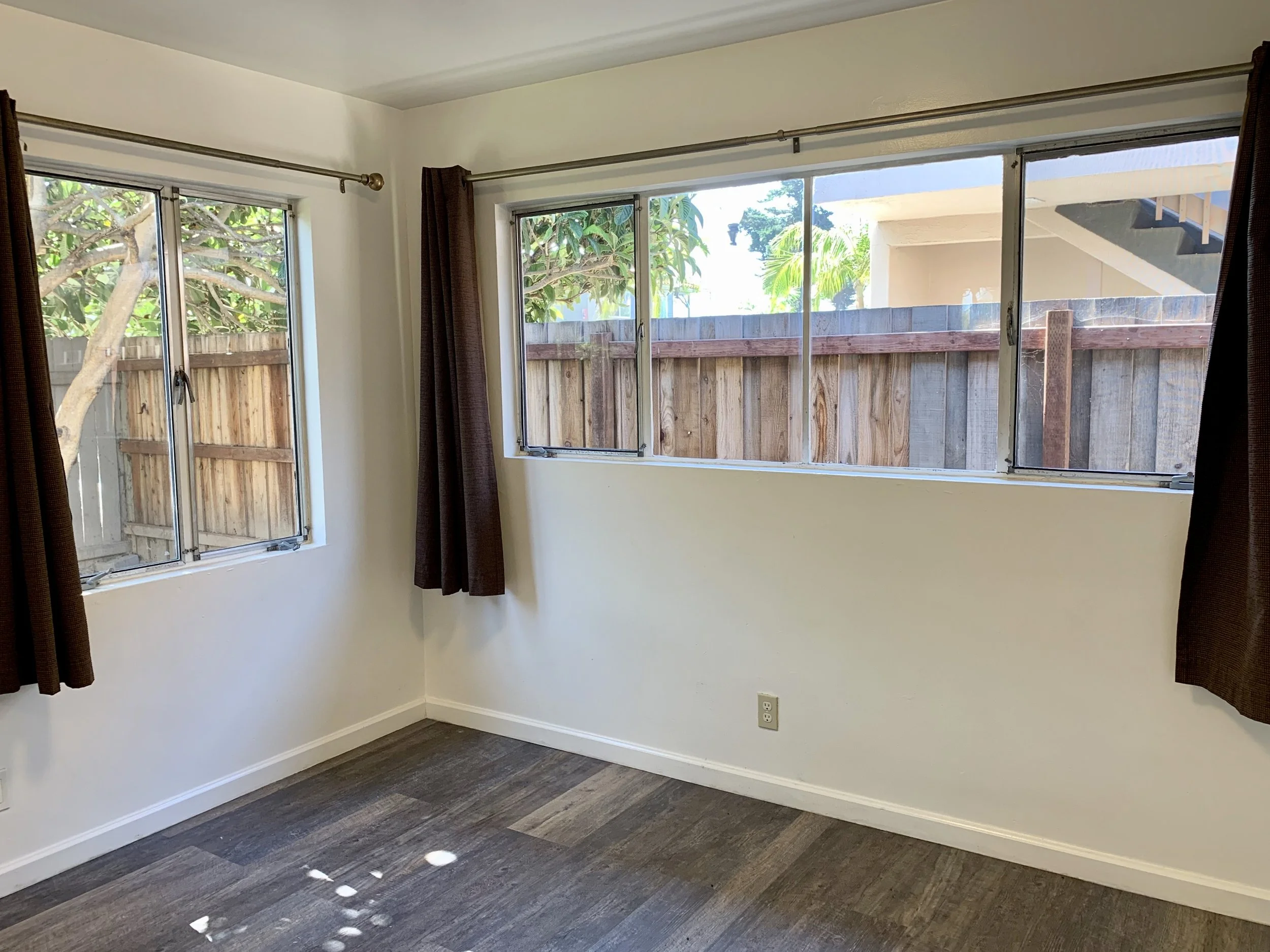Empty room with white walls, dark hardwood flooring, two large windows with brown curtains, and a wooden fence outside. Sunlight creates circular light spots on the floor.
