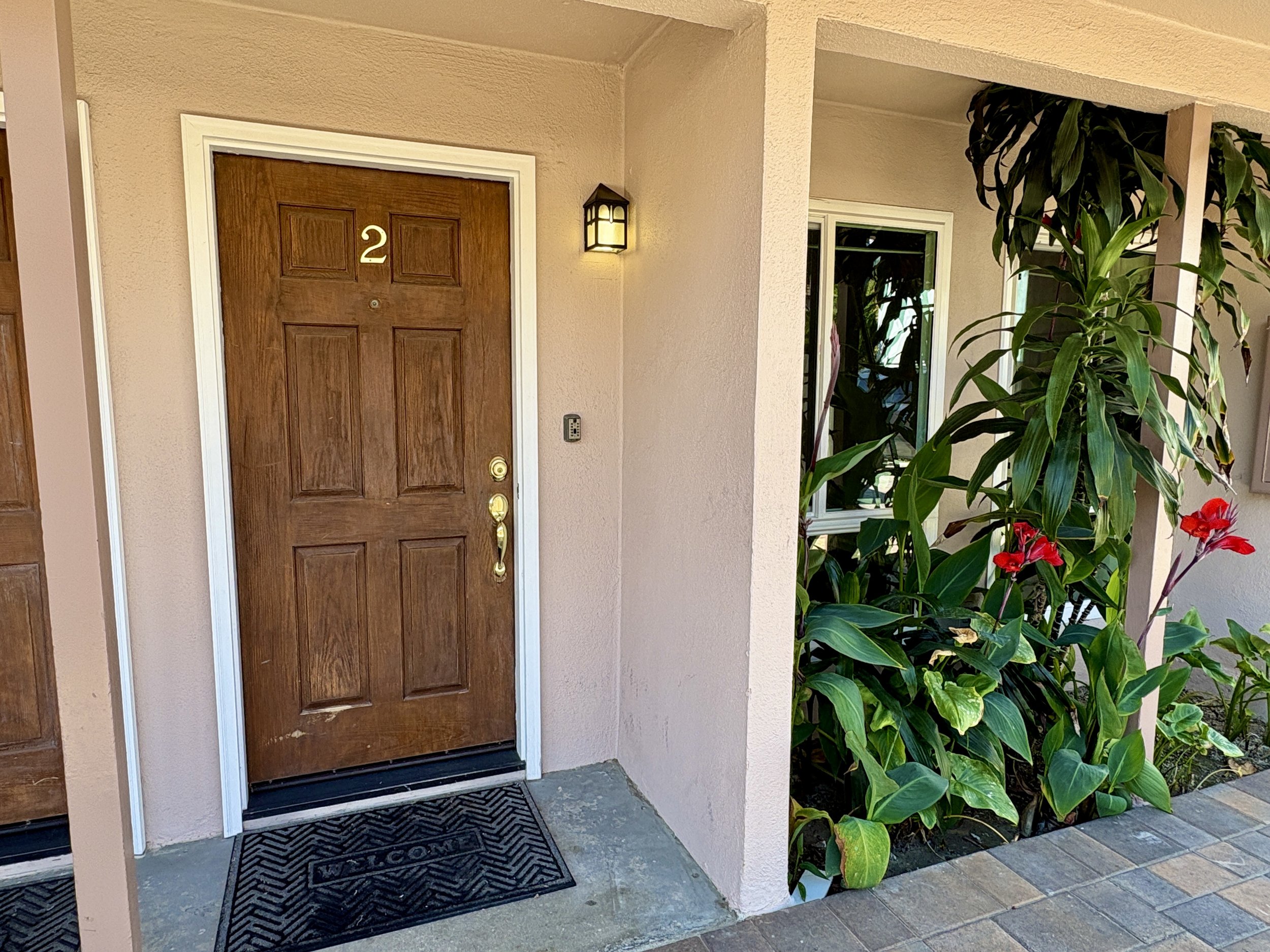 The image shows the front door of a house with the number 2. The door is made of wood, and there is a black doormat in front of it with the word "Welcome". To the right of the door, there are large green plants with red flowers, and a window is visible in the background.