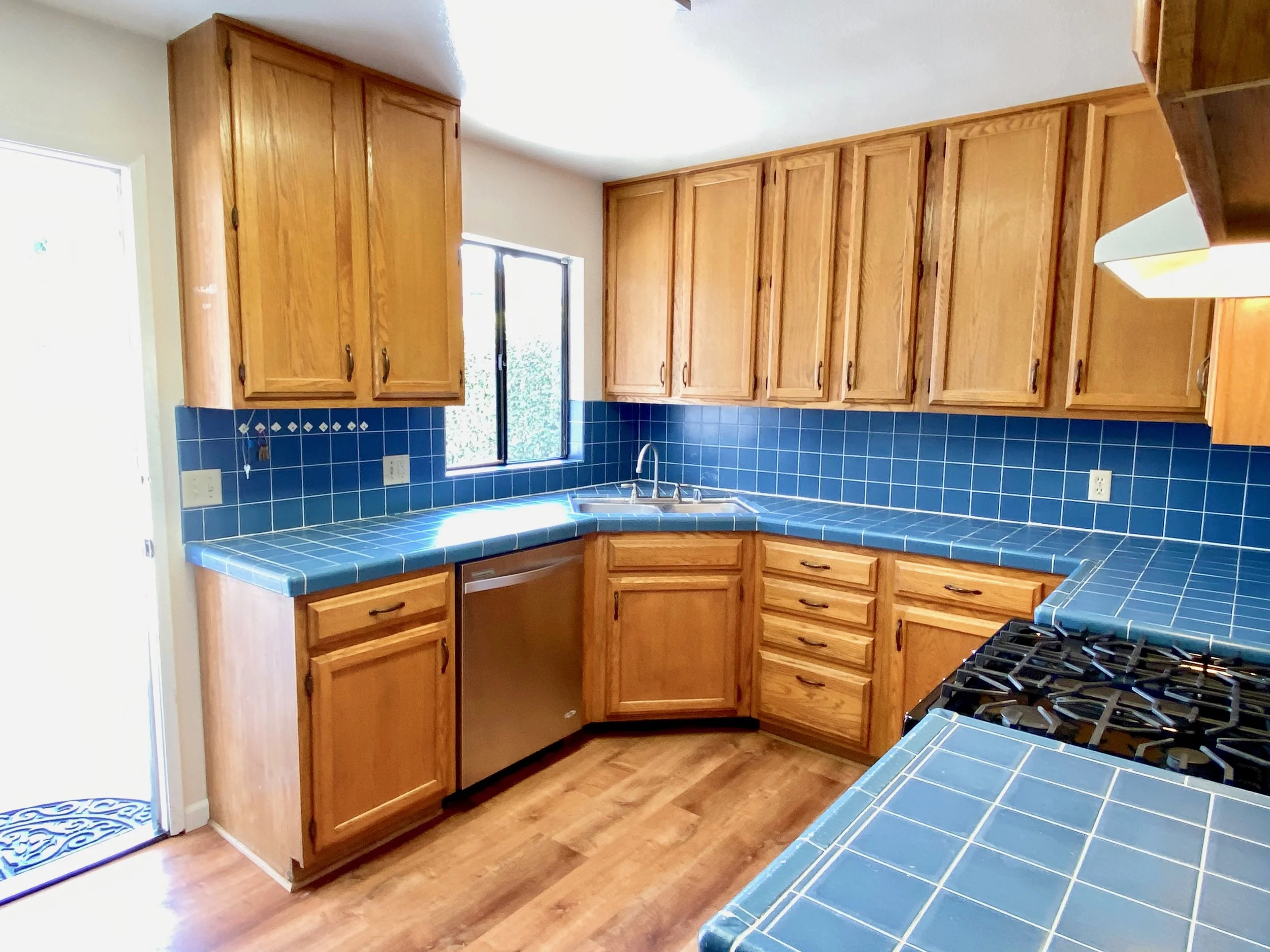 A kitchen with wooden cabinets, blue tiled countertops, a window above the sink, and a dog door in the door to the outside.