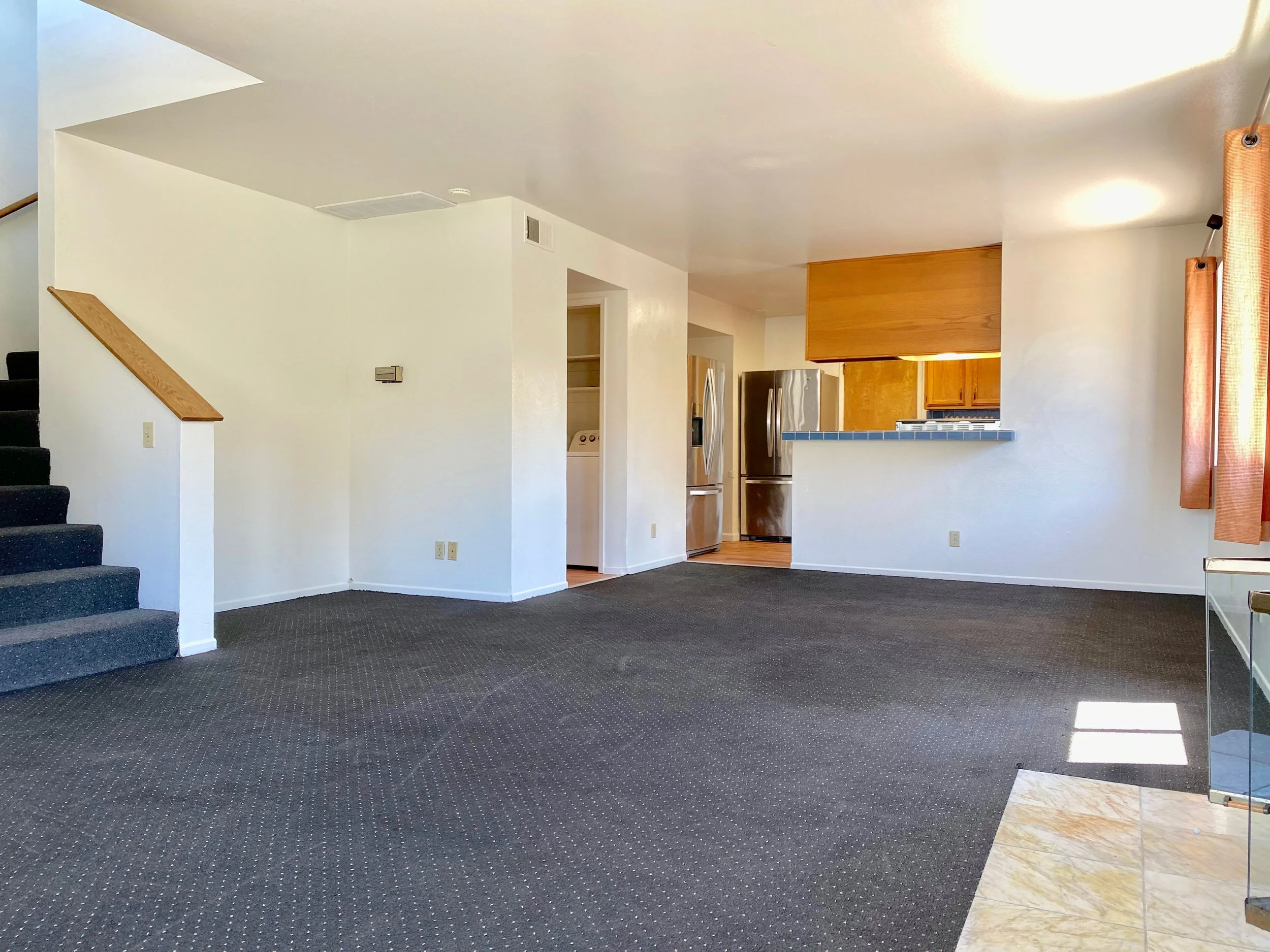 Interior view of a living space with a staircase on the left, a partial kitchen in the background, and a large window with curtains on the right. The room has carpeted flooring and minimal furnishings.