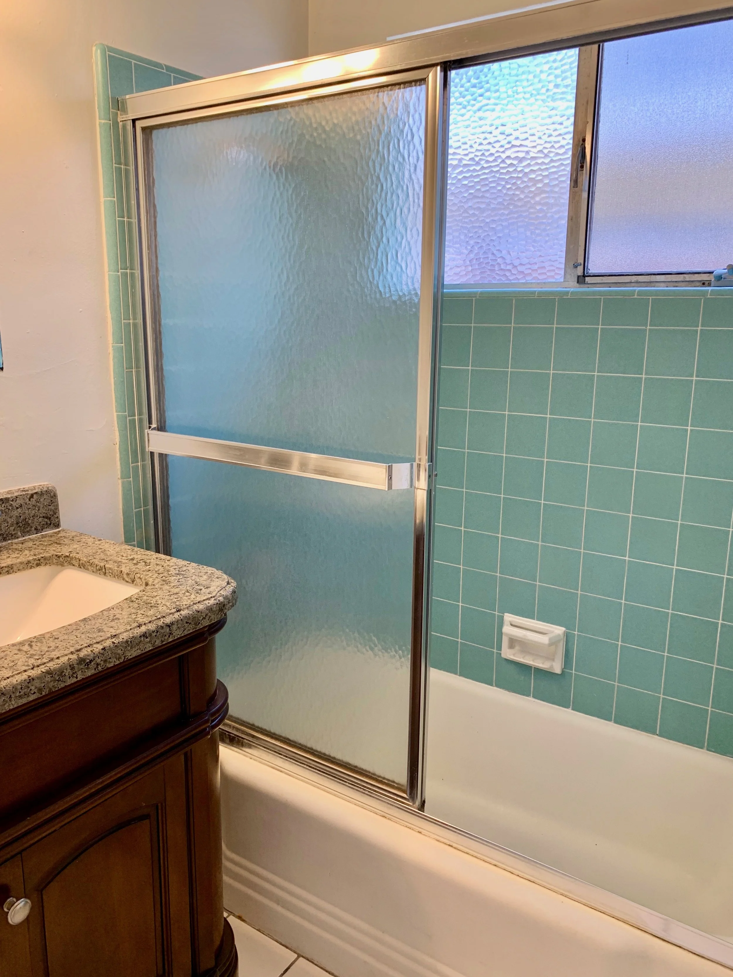 A bathroom with a wooden vanity, a granite countertop, and a white sink. There's a frosted glass shower with a metal frame and teal wall tiles inside. A frosted glass window provides natural light.