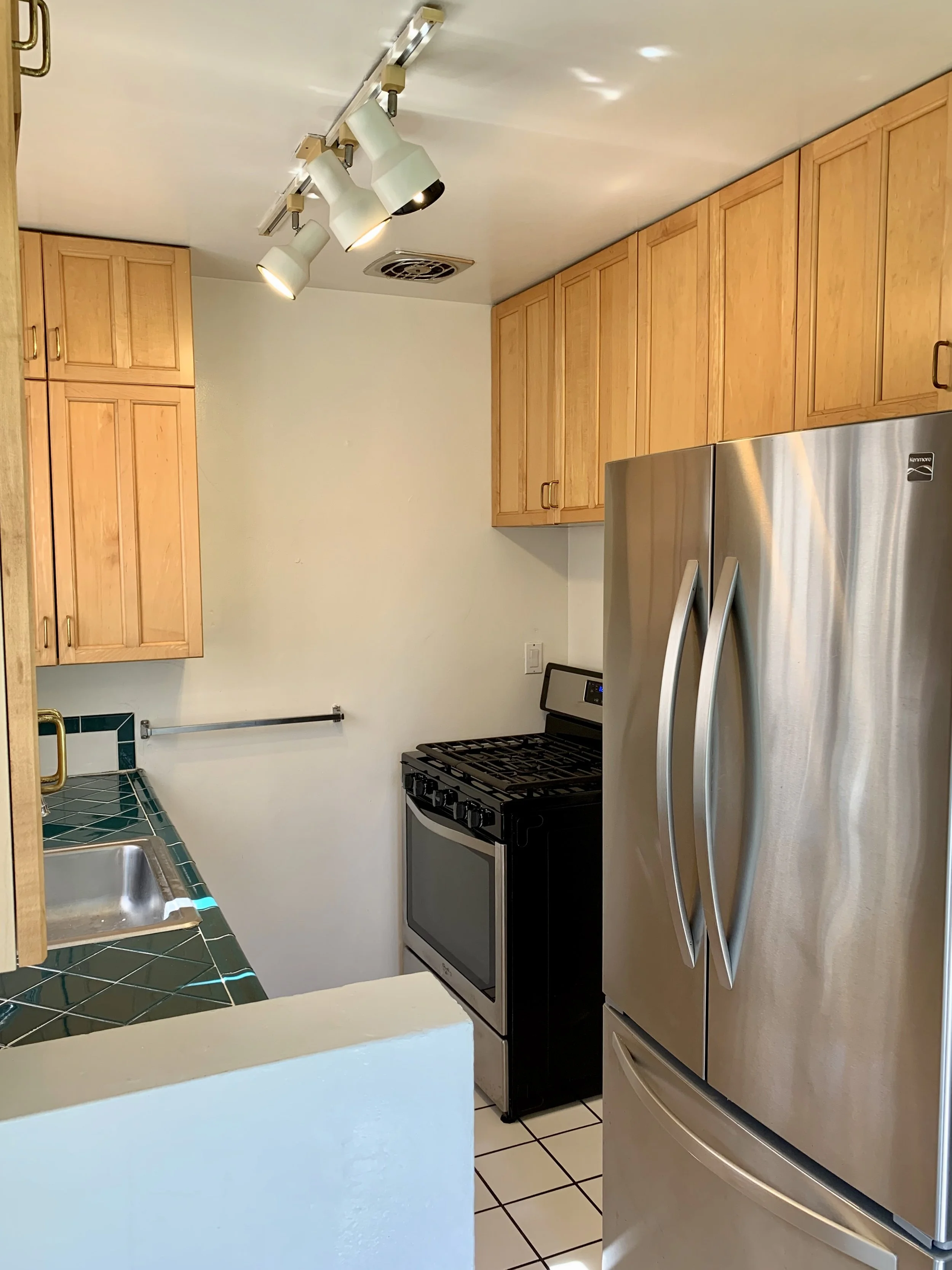 A kitchen with wooden cabinets, a stainless steel refrigerator, a black stove, a sink with a green tiled countertop, and a ceiling light fixture.