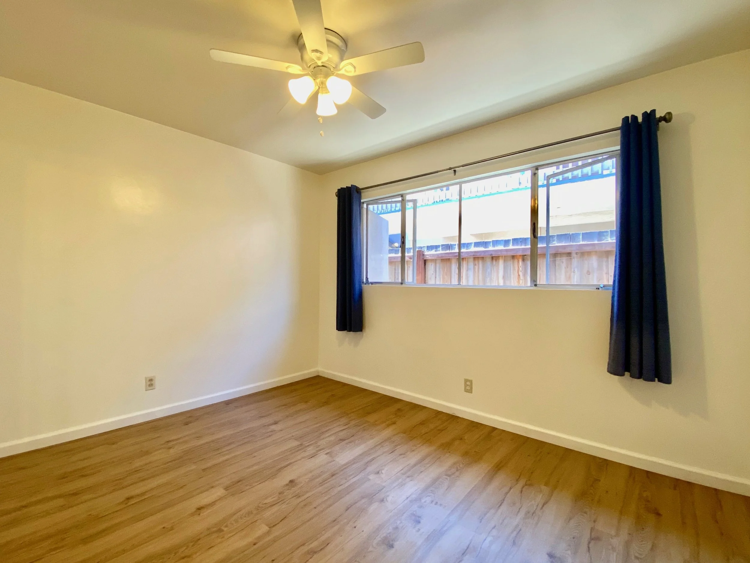 Empty room with beige walls, wooden floor, a window with blue curtains, and a ceiling fan with lights.