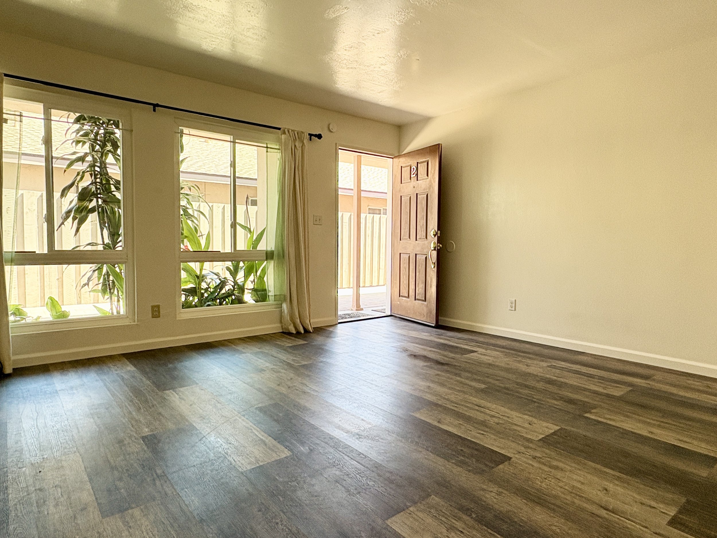 An empty living room with wood-look flooring, large windows with a plant outside, and an open front door leading to a paved driveway.