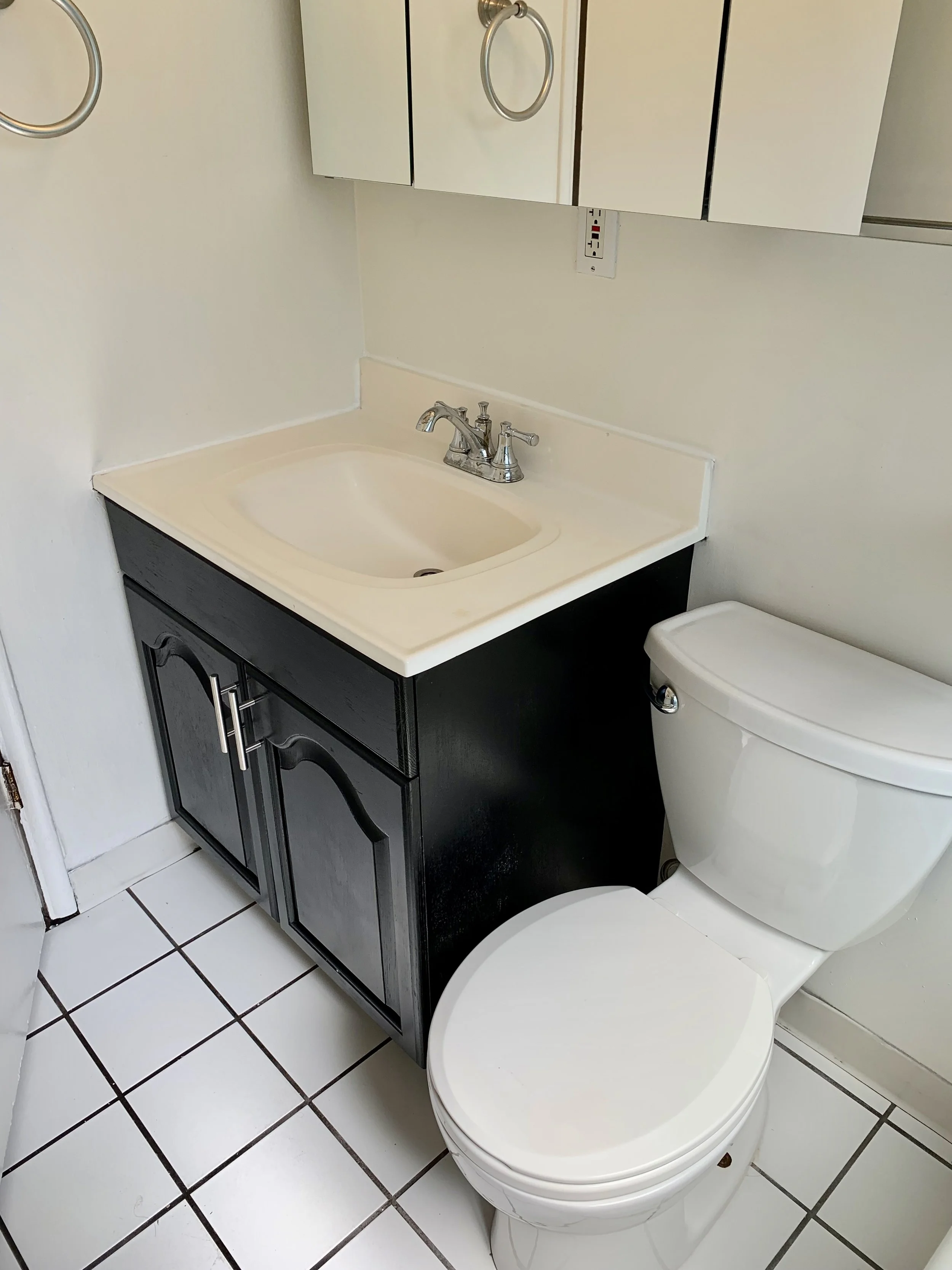 Bathroom with a white toilet, a sink with a black cabinet, a mirror cabinet above the sink, tile flooring, and electrical outlets on the wall.
