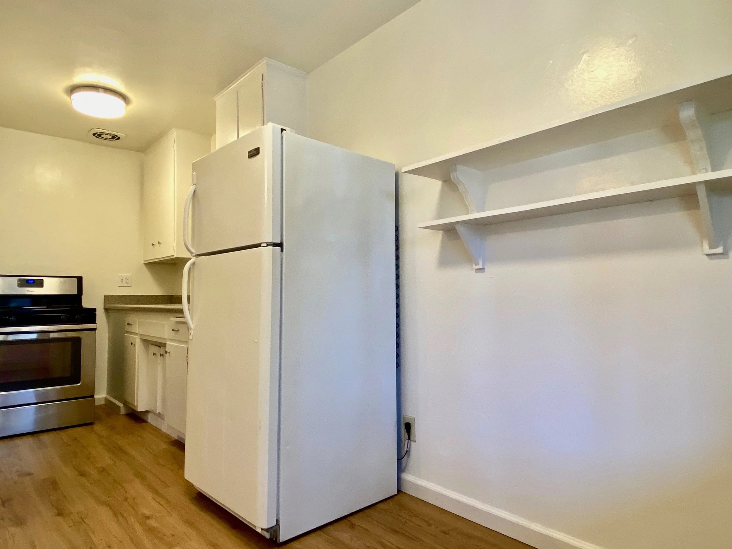 A partial view of a kitchen with a white refrigerator, white cabinets, shelving, a stove, and hardwood floors.