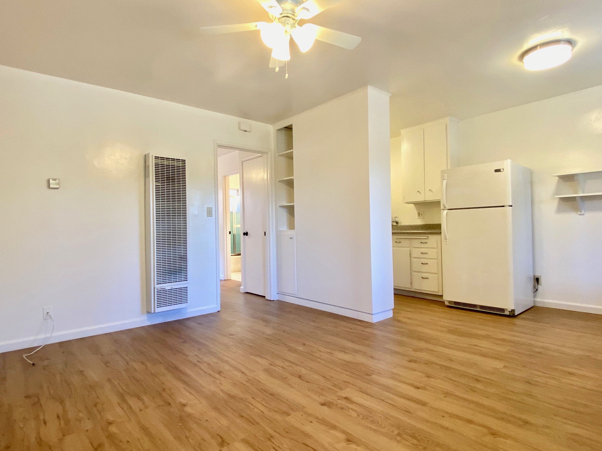 Empty living room with hardwood floors, white walls, ceiling fan, and partial view of kitchen with white refrigerator and cabinets.