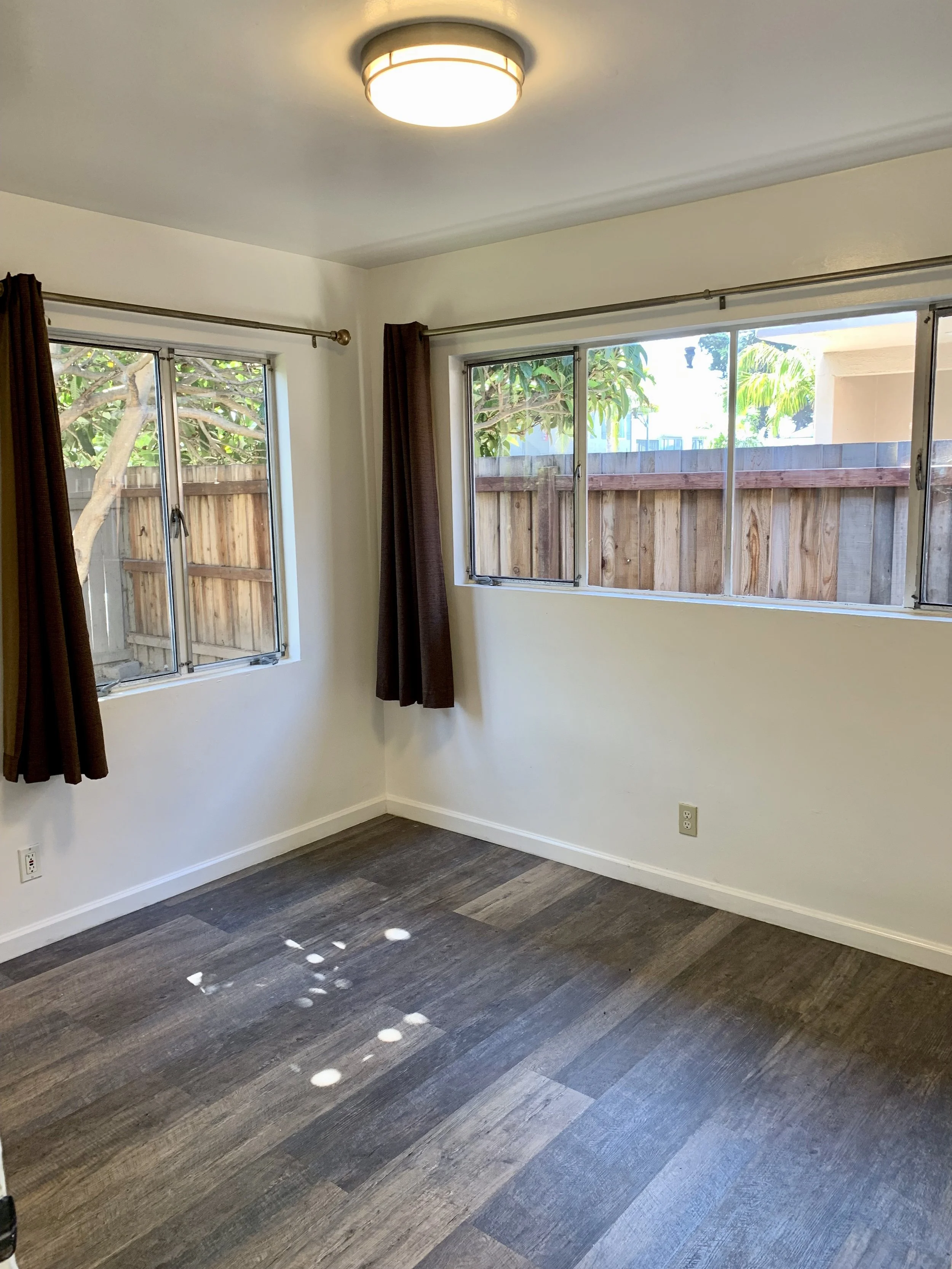 Empty room with hardwood flooring, white walls, a ceiling light, and three windows with brown curtains showing a wooden fence and green trees outside.