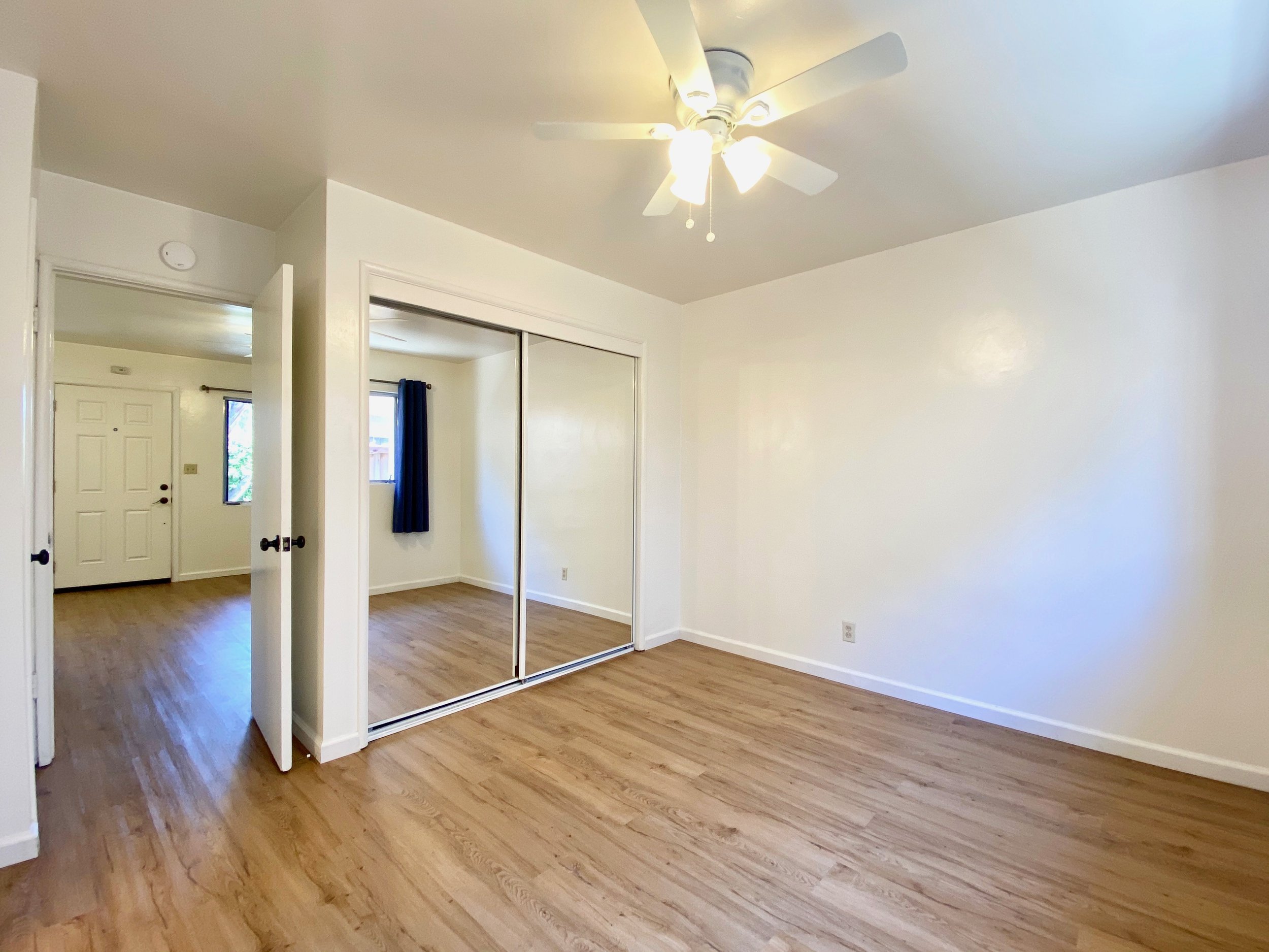 A room with wooden floors, white walls, a ceiling fan with lights, a closet with mirrored sliding doors, a window with dark curtains, and a door to the living room.