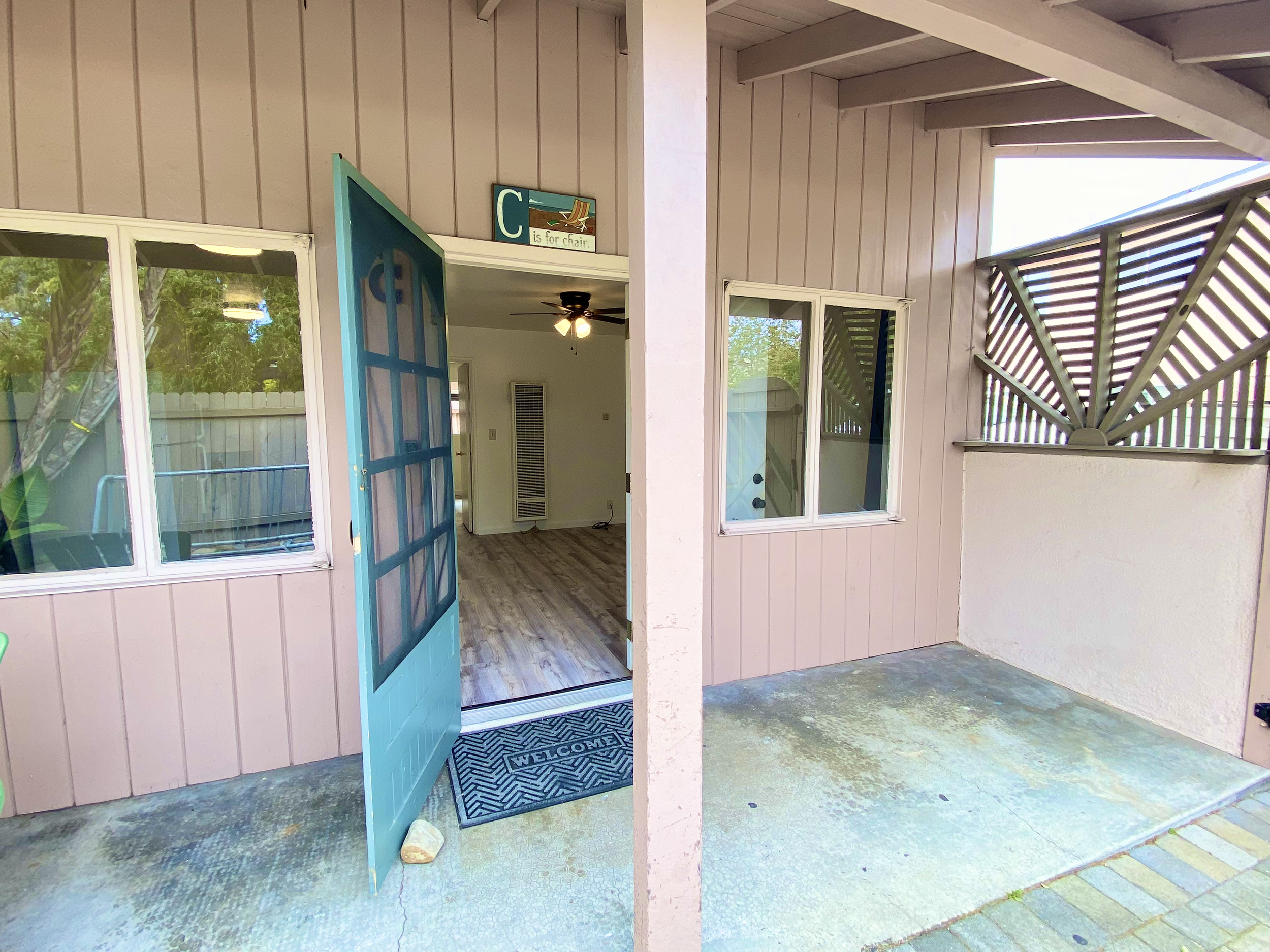 Open blue front door of a house, with a welcome mat outside, leading into a room with a ceiling fan, and windows. Part of the exterior porch with concrete flooring and siding visible.