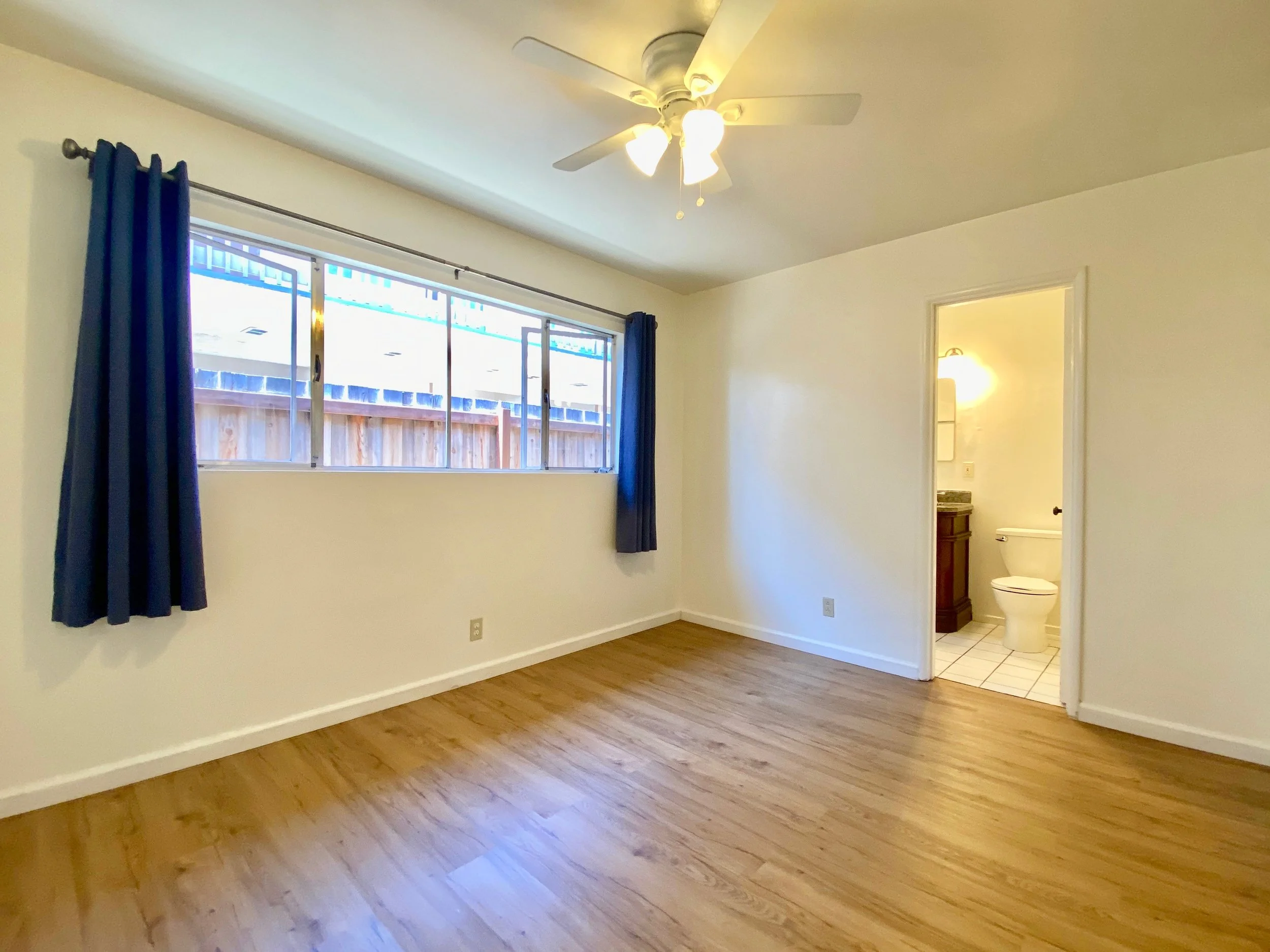 Empty room with hardwood floors, large window with blue curtains, ceiling fan with lights, and a doorway leading to a small bathroom with a toilet inside.