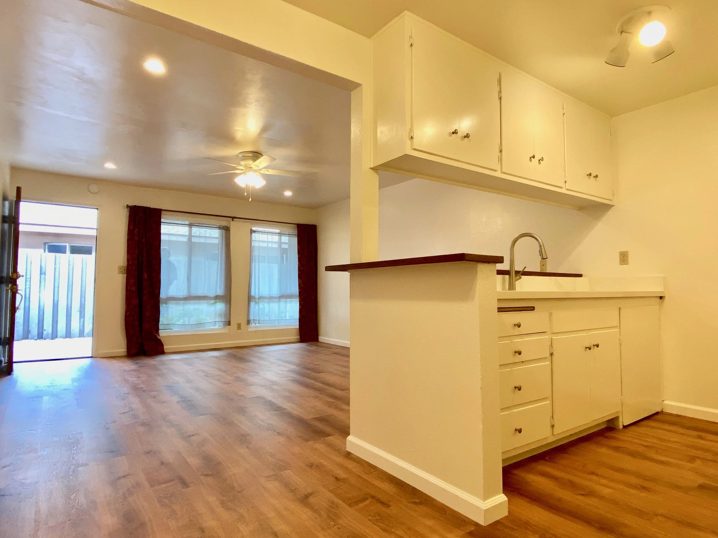 An open-plan living area with hardwood floors, large windows with curtains, a ceiling fan, and an adjoining kitchen with white cabinets and a sink.