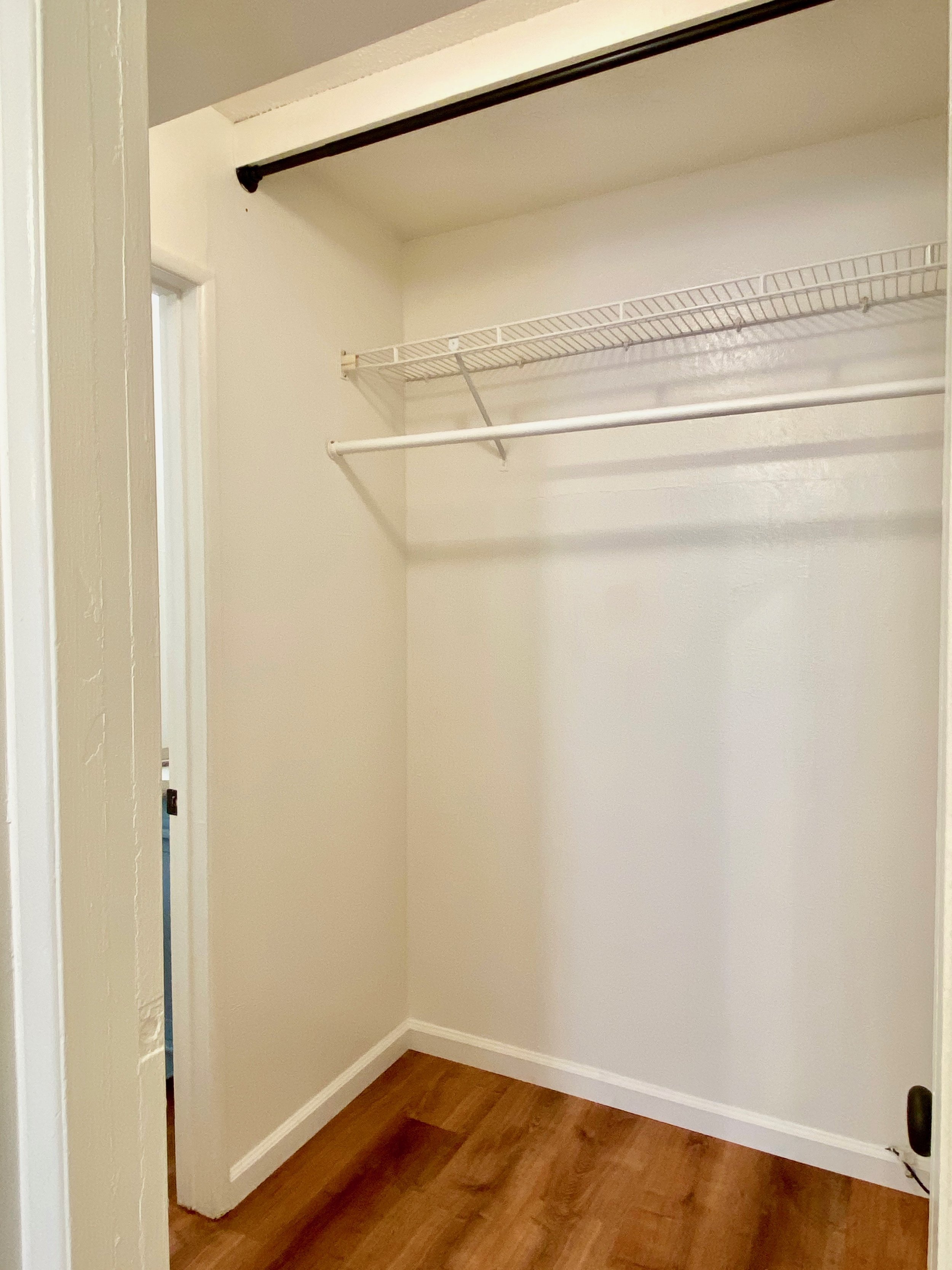 Empty closet with wire shelf and clothes rod, hardwood floor, white walls, visible doorway, and an electrical outlet on the right wall.